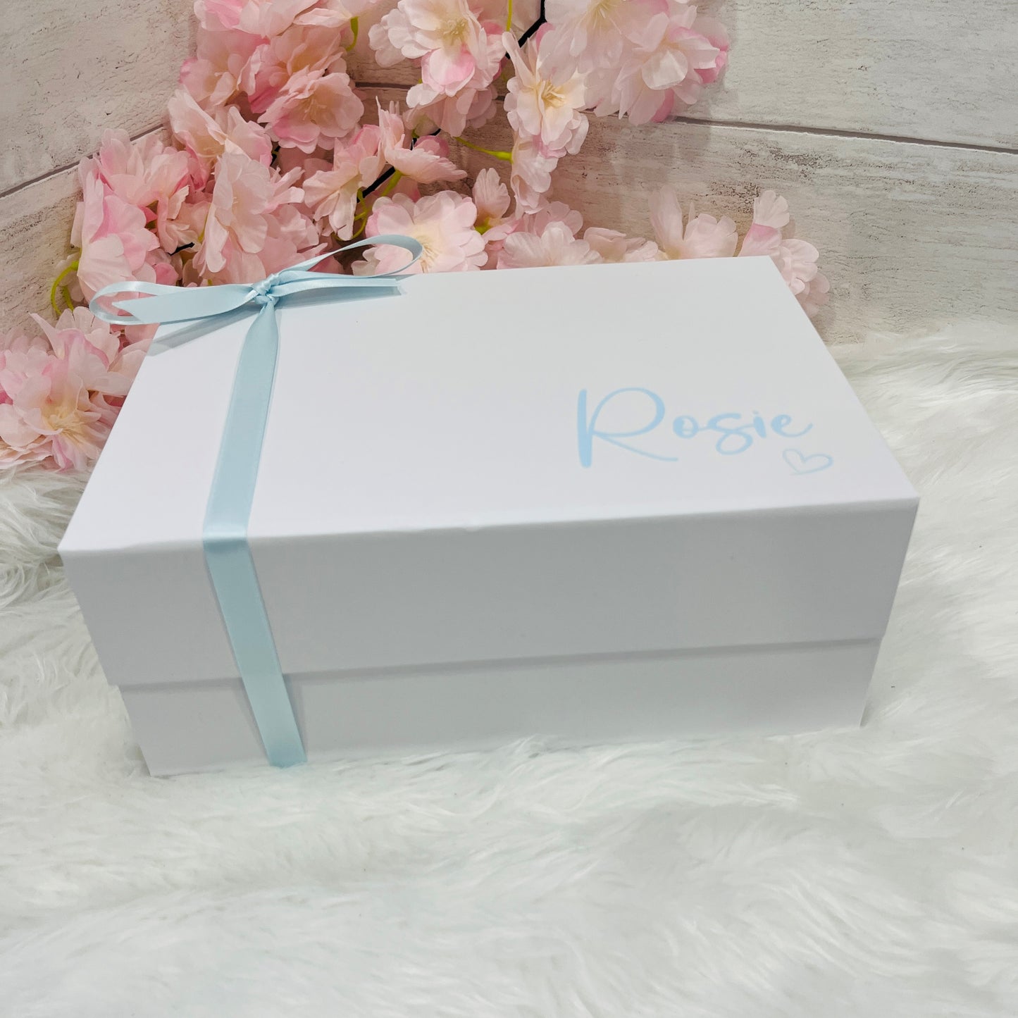 White gift box with a blue ribbon and 'Rosie' inscription, placed on a white surface with pink flowers in the background.