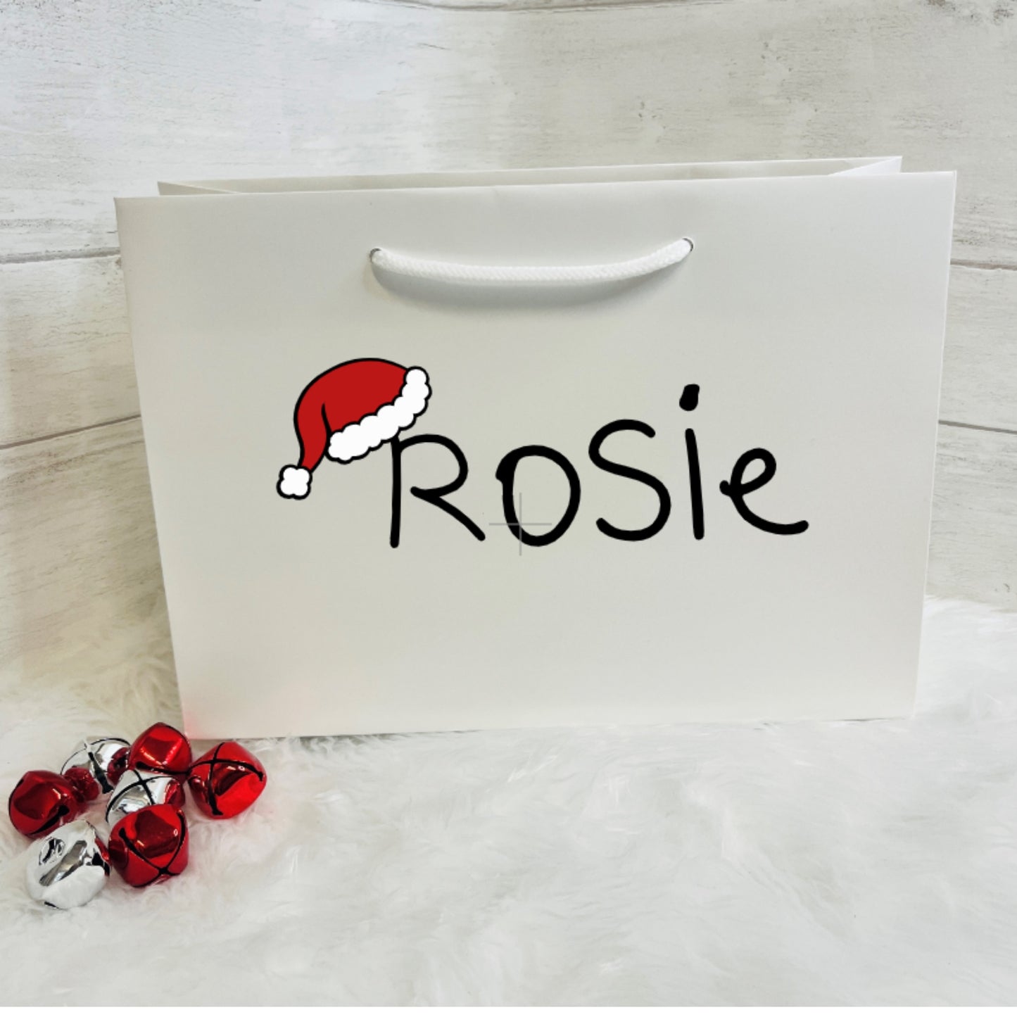 White gift bag with 'Rosie' and a Santa hat design, accompanied by red and silver bells on a light wooden surface.