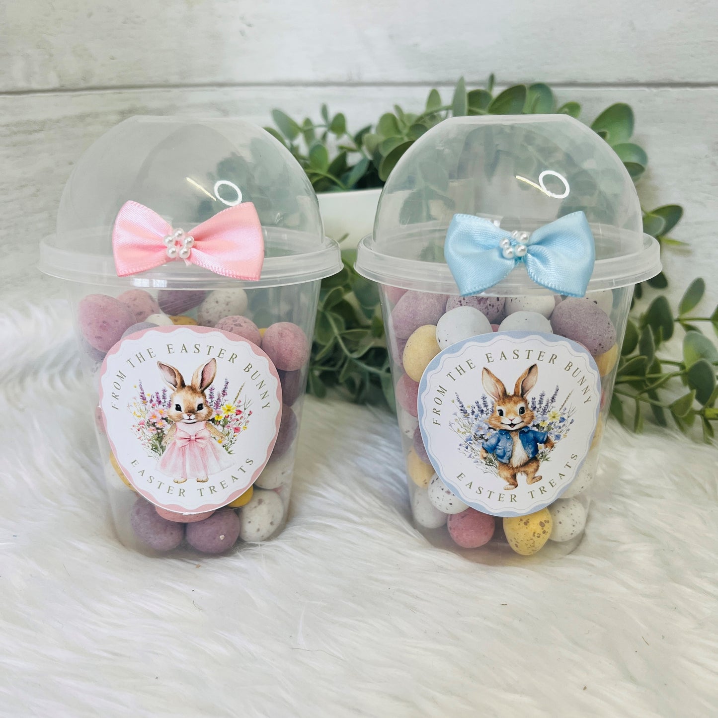 A pair of Easter Dome Snack Pots featuring a plastic cup with a blue and pink bunny wreath design and a blue and pink bow on the lid, filled with various small snacks.