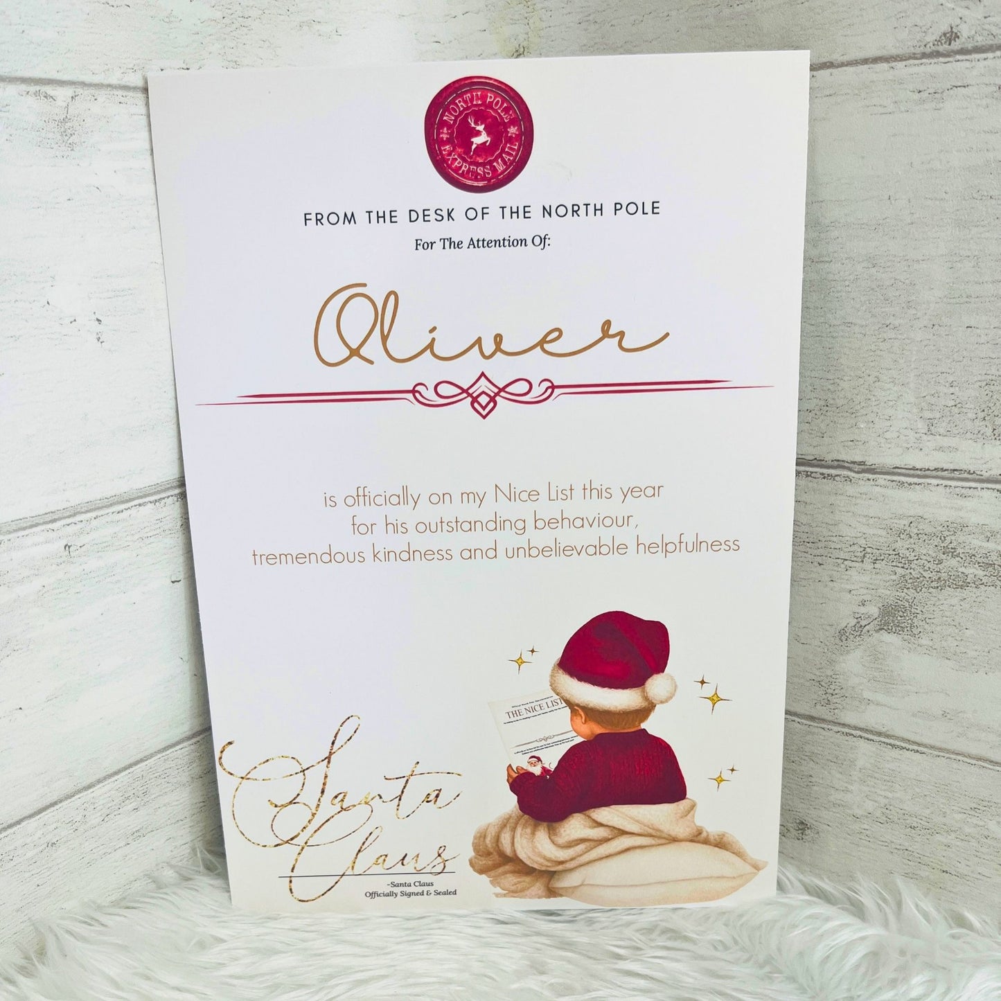 An A4 printed Nice list card addressed to 'Oliver' from the North Pole, featuring gold cursive text, a red circular logo, and an illustration of a young boy in a red jumper wearing a santa hat. The card is photographed standing upright against a light gray wood-plank background with a white faux-fur surface underneath.