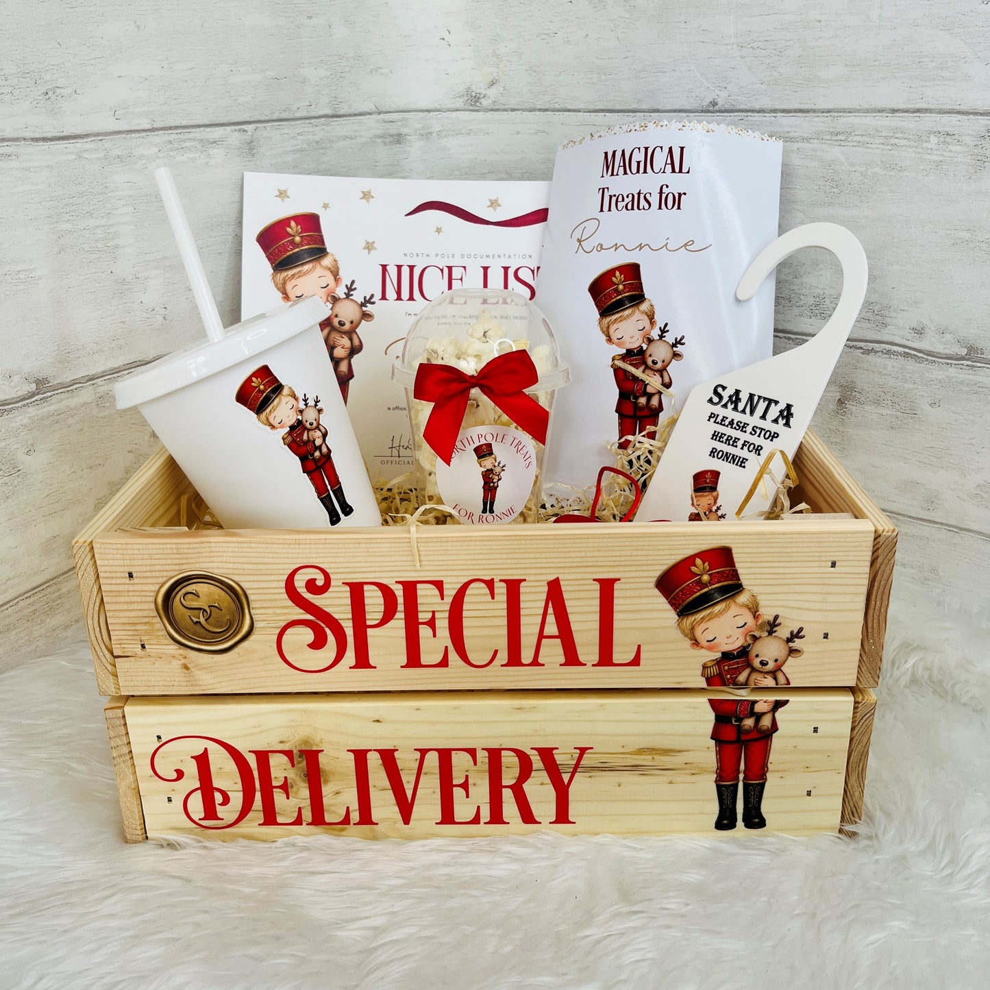 A Christmas-themed gift basket set in a wooden crate labeled 'SPECIAL DELIVERY,' filled with holiday treats including a cup, packaged snacks, a small ornament, and printed cards featuring nutcracker illustrations and the text 'MAGICAL Treats for Ronnie' and 'Santa Please Stop here for Ronnie'.