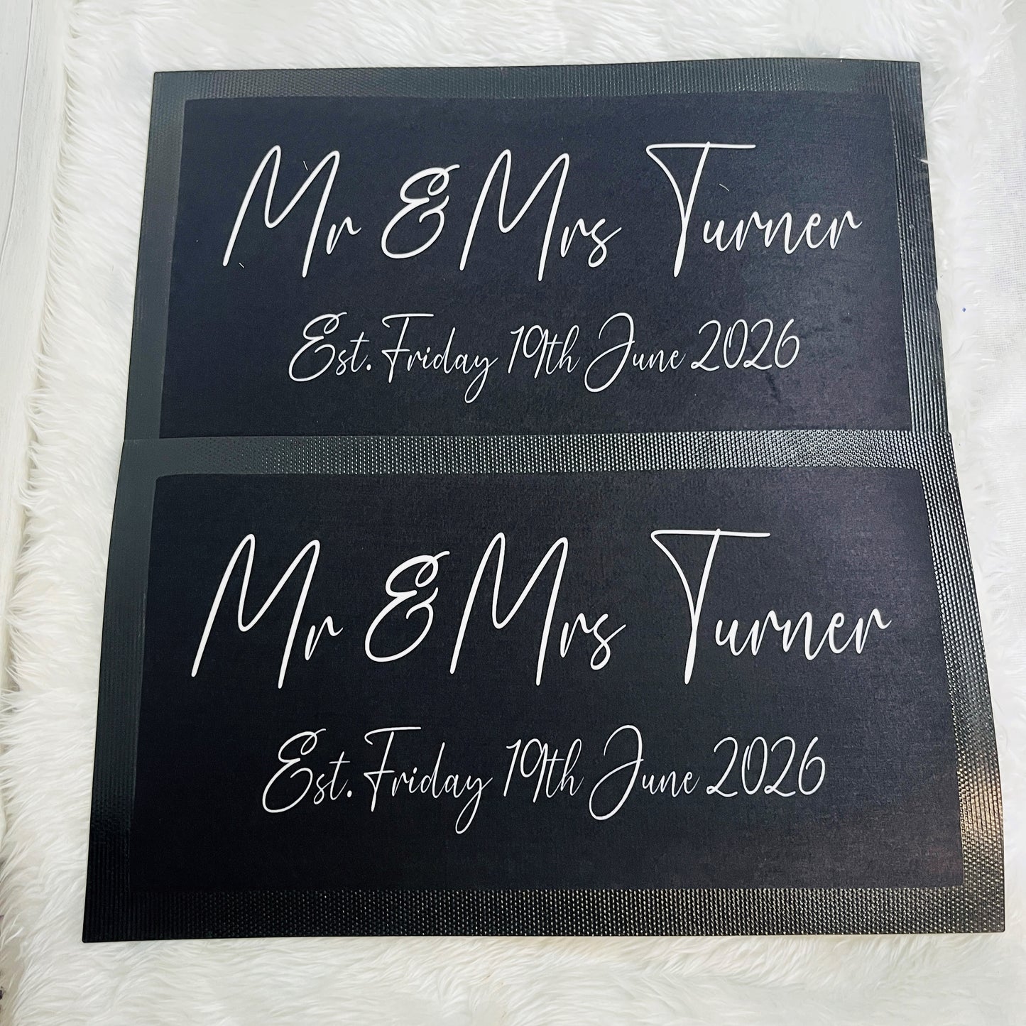 Two matching black rectangular beer runners with white script text reading 'Mr & Mrs Turner' and 'Est. Friday 19th June 2026', placed on a white furry surface.