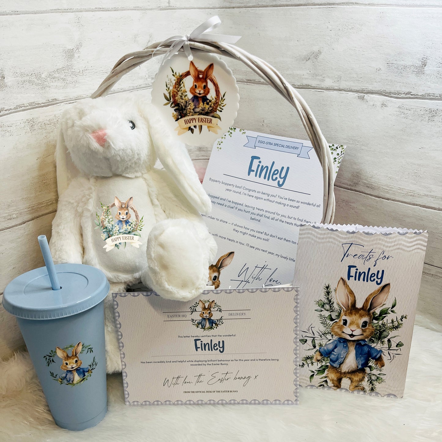 Blue Easter gift basket with plush bunny, tumbler, and personalized cards on a white fluffy surface.