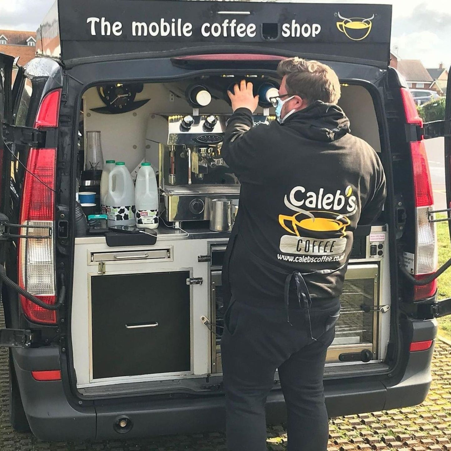 A person wearing a black hoodie with 'Caleb's Coffee' branding on the back, standing near a vehicle.