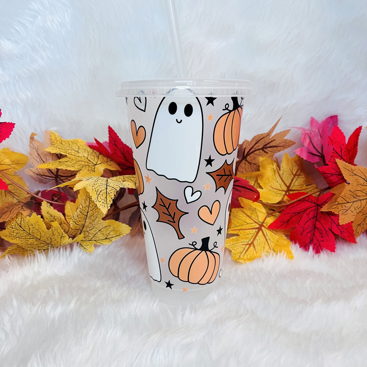 A frosted cold cup tumbler with a lid and straw, decorated with a cute Halloween pattern of white ghosts, orange pumpkins, brown leaves, and orange hearts. The cup is set on a white faux fur surface and surrounded by colorful autumn leaves.
