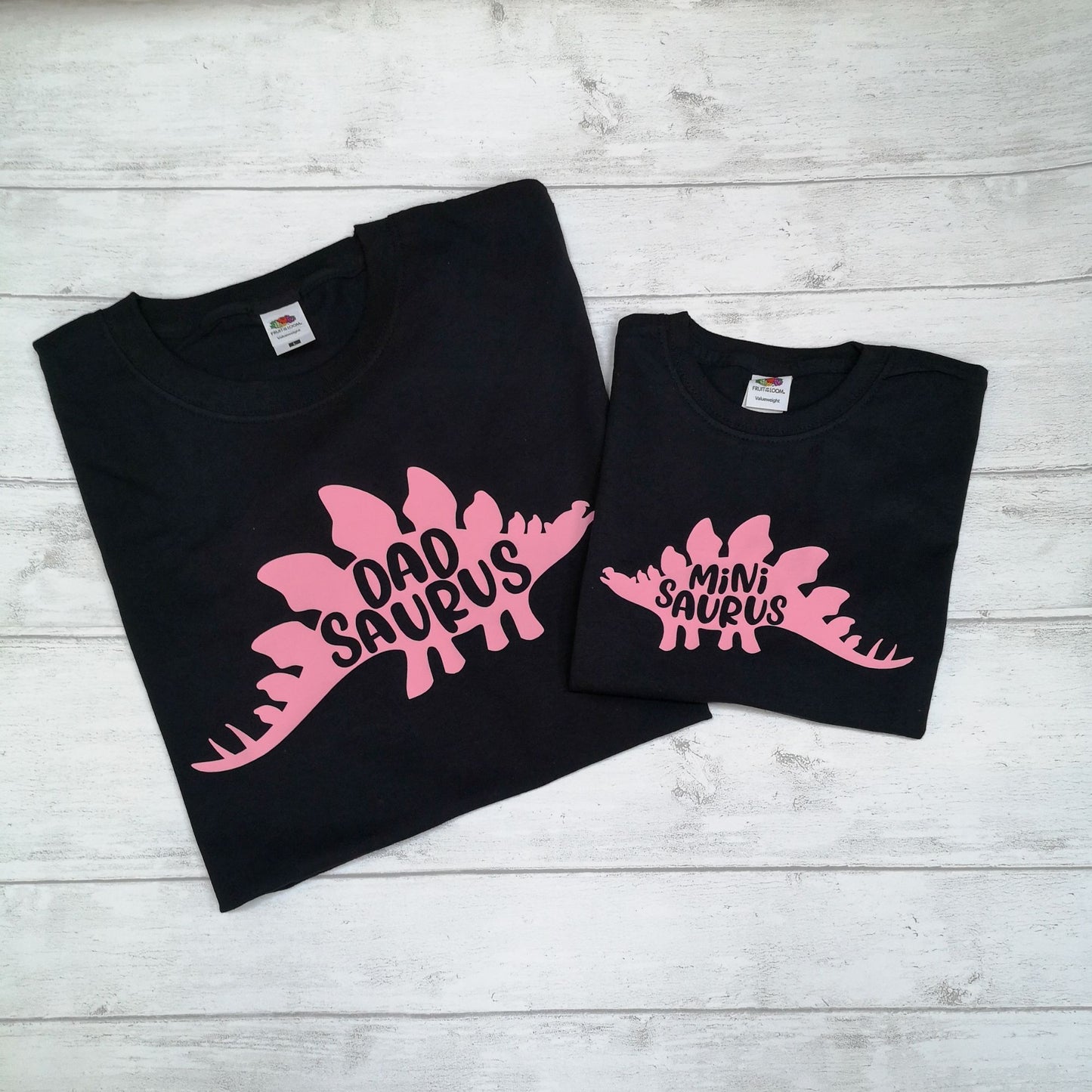 Two black sweatshirts with pink dinosaur designs on a light wooden surface