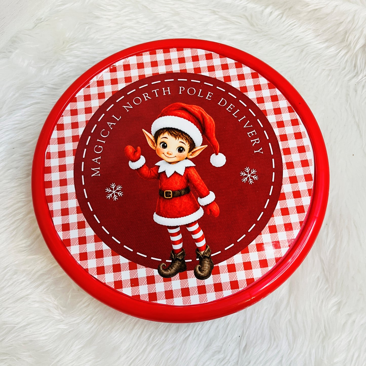 A round red and white gingham sweet tin featuring a cartoon elf in a Santa outfit with the text 'Magical North Pole Delivery' on a white faux fur background.