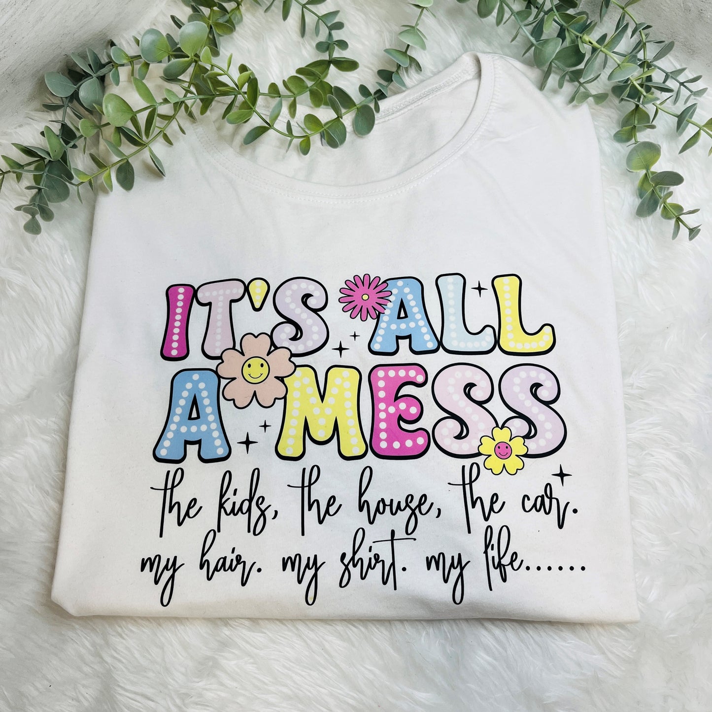 White crew neck T-shirt with the phrase 'It's all a mess' printed in colorful letters, surrounded by illustrations of flowers