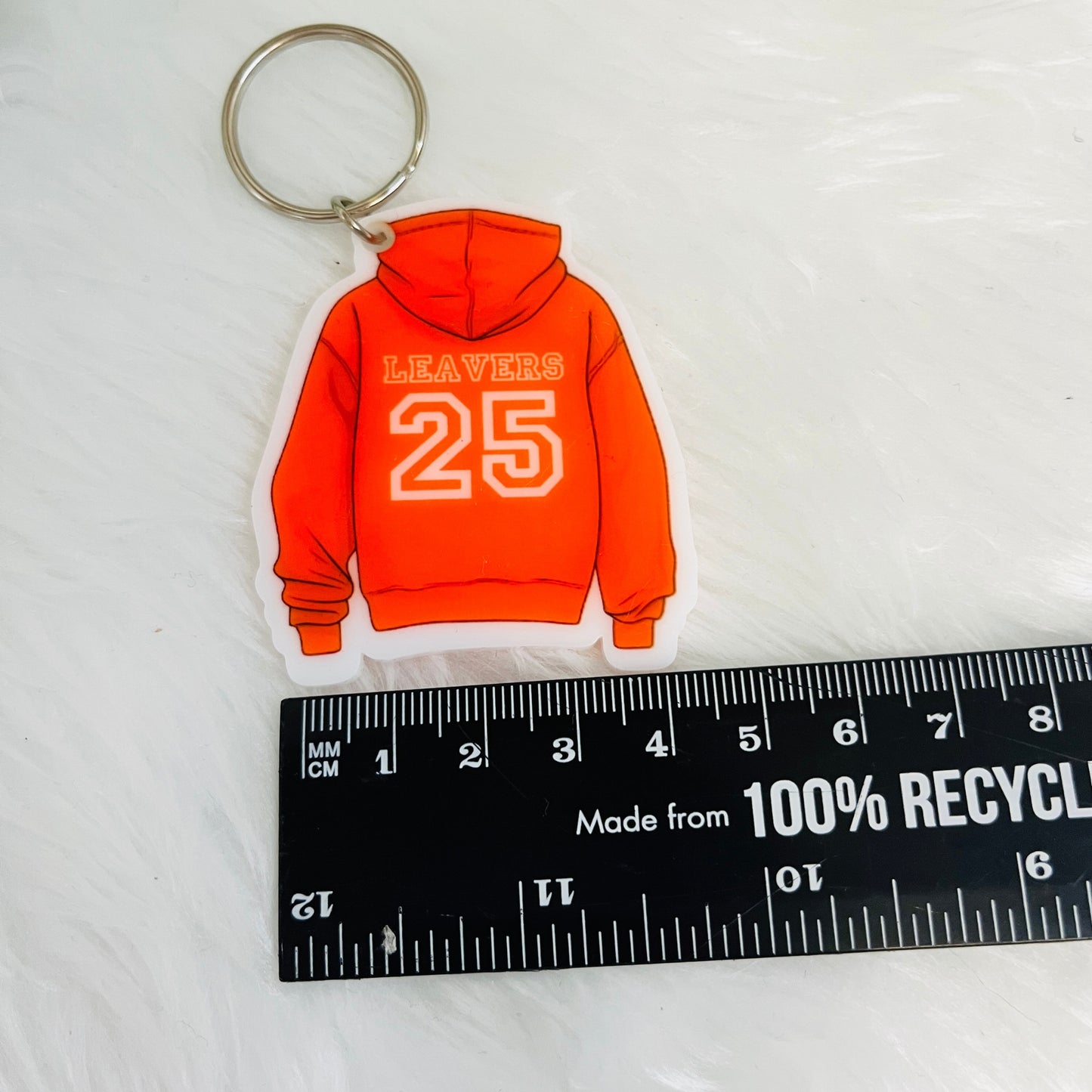 Keychain shaped like an orange hoodie with 'LEAVERS 25' text, next to a ruler for scale on a white background.