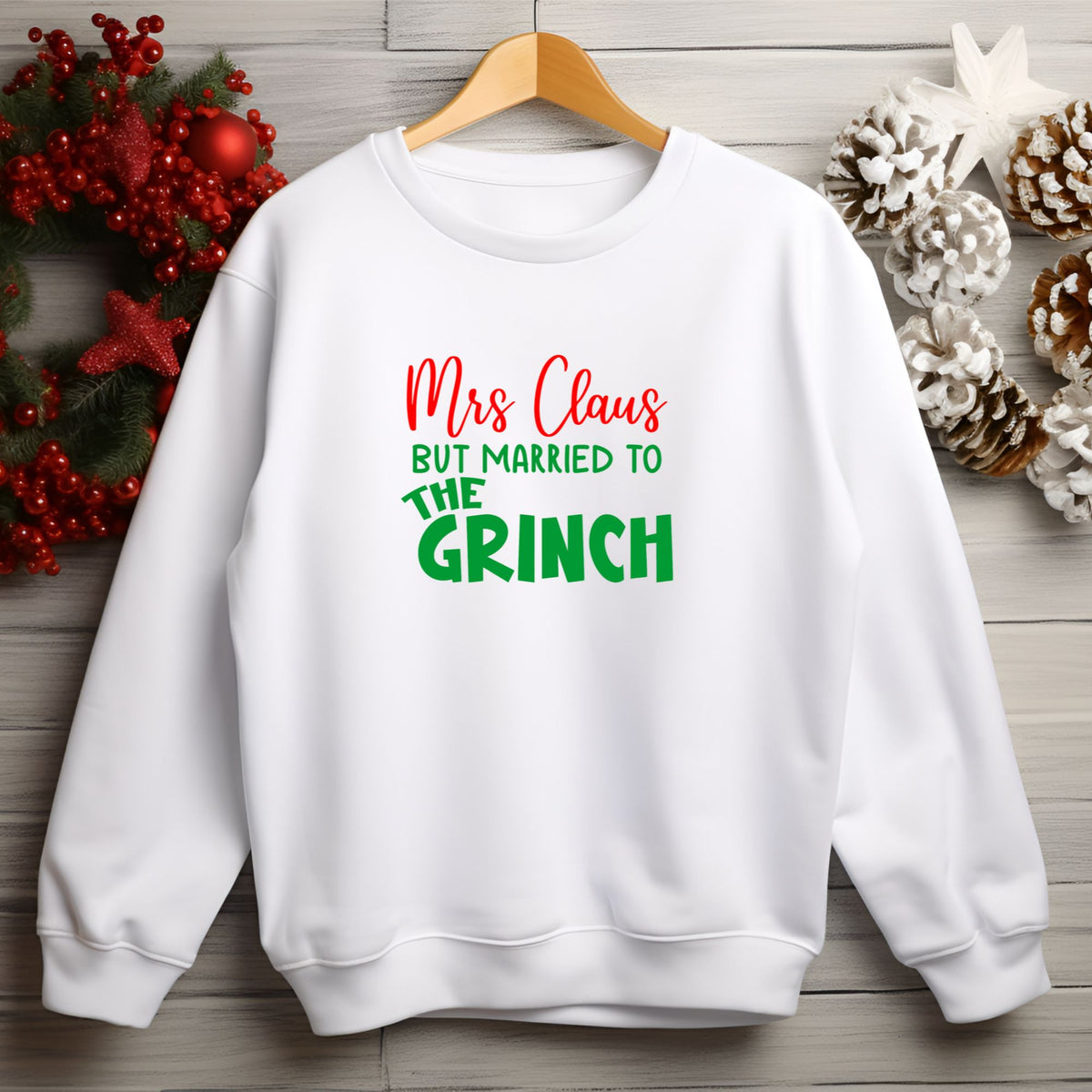 A white crewneck sweatshirt hanging on a wooden hanger against a light gray wood plank background, decorated with Christmas wreath and pinecones. The sweatshirt features the text 'Mrs Claus BUT MARRIED TO THE GRINCH' in red and green lettering.