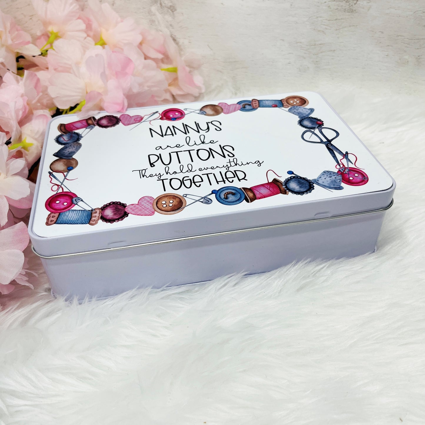 A white metal tin laying face down to show depth of tine with 'nannys are like buttons, they hold everything toether' on the lid with a sewing themed design around the edge
