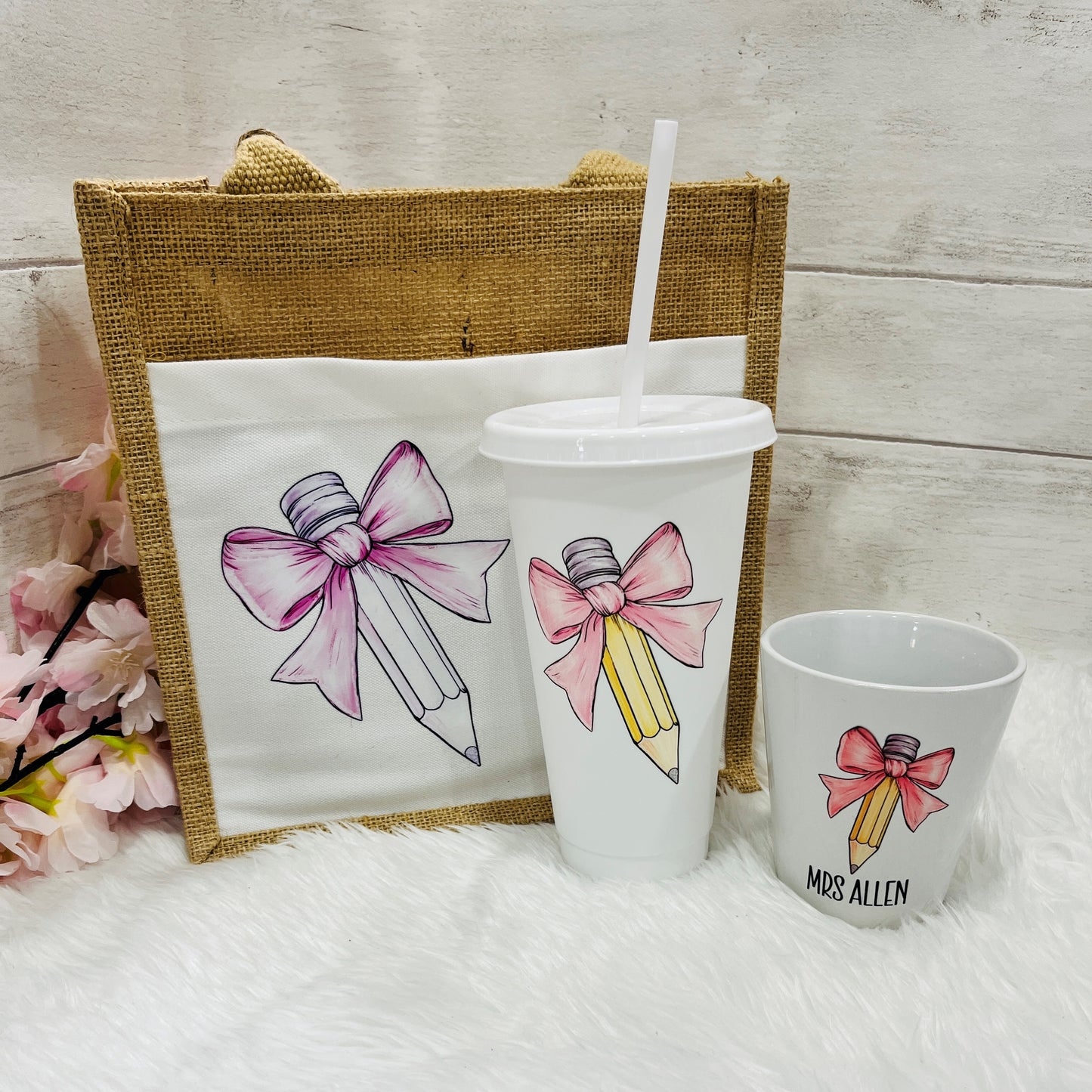 A set of personalized teacher's gifts including a jute bag with a bow and pencil design, a white cold cup with a bow and a pencil pot with pale pink bow design.