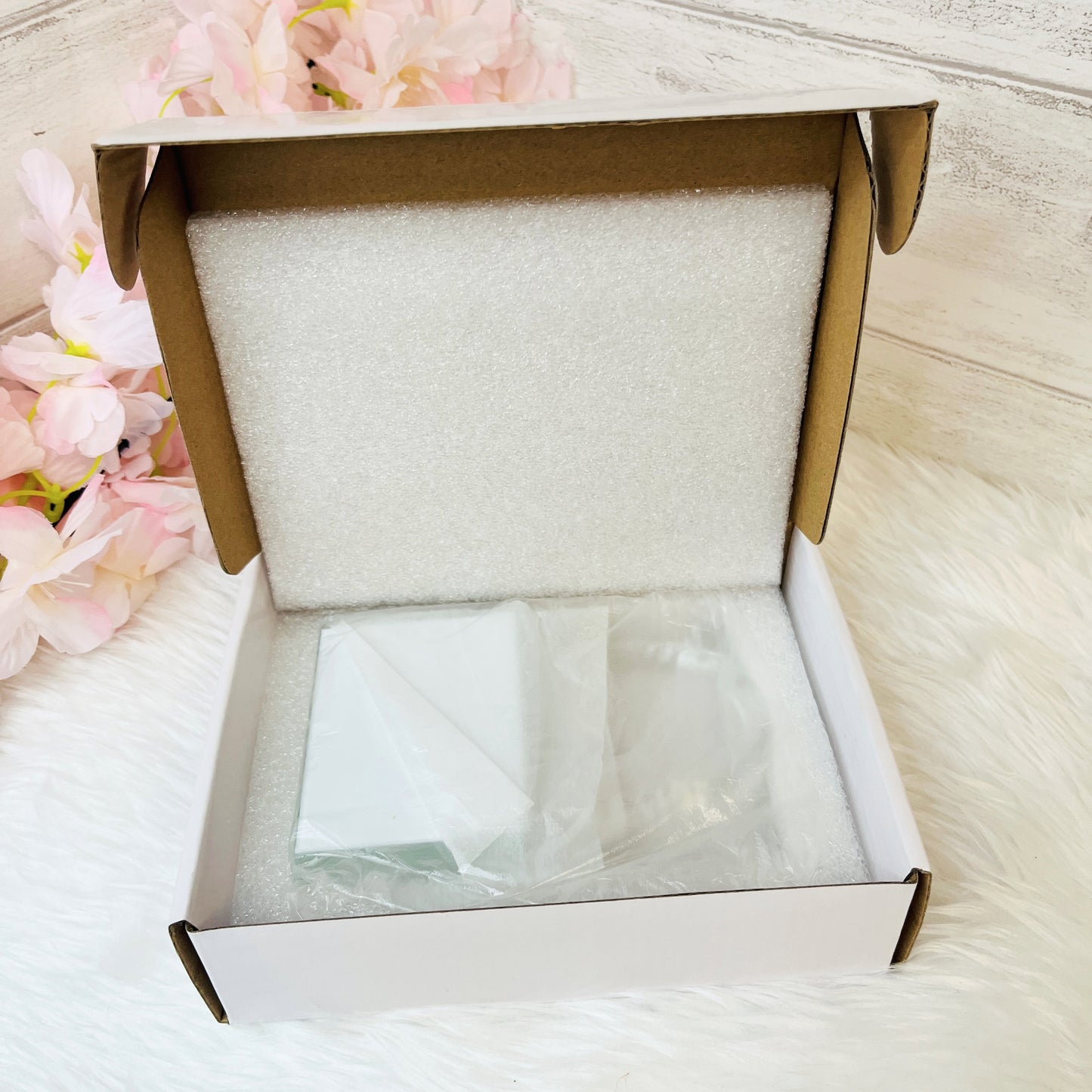 An open white cardboard box with a white foam insert, containing a crystal love lock, placed on a white furry surface with pink flowers in the background.