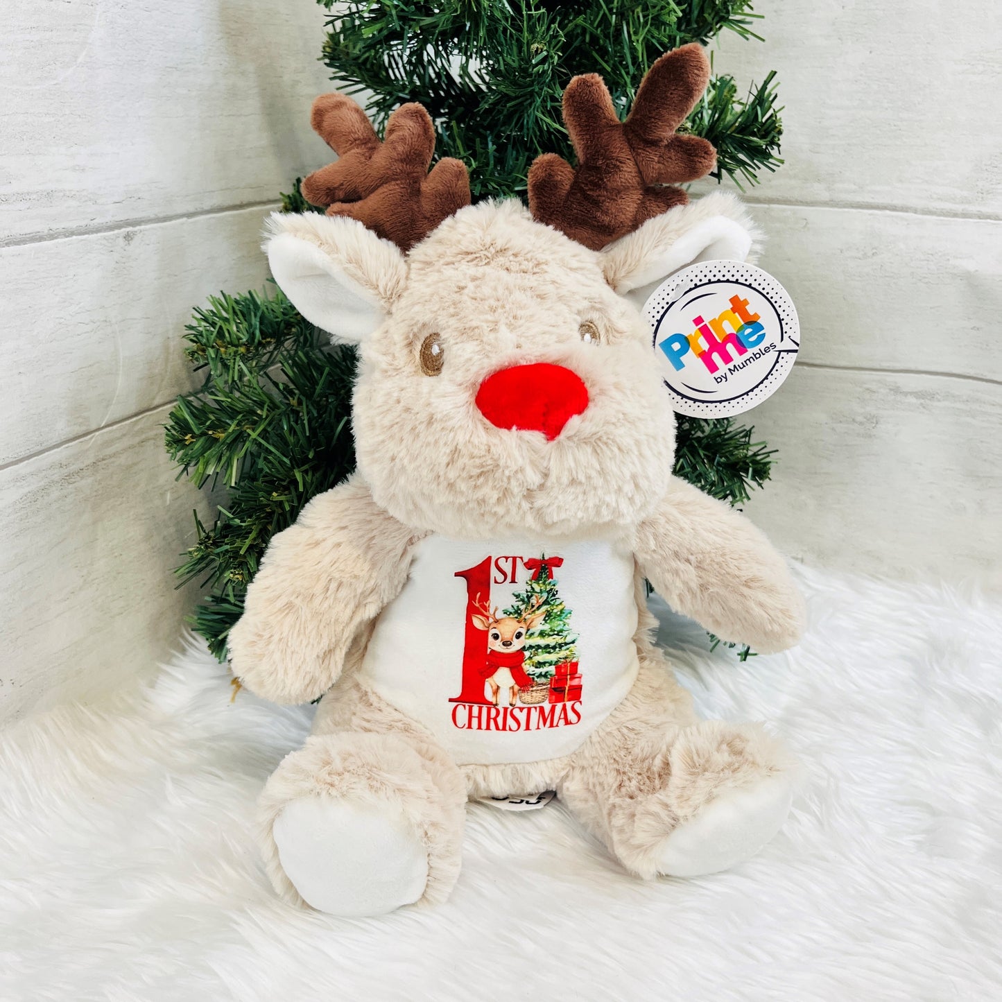 Plush reindeer toy with 'First Christmas' message in front of a small Christmas tree.