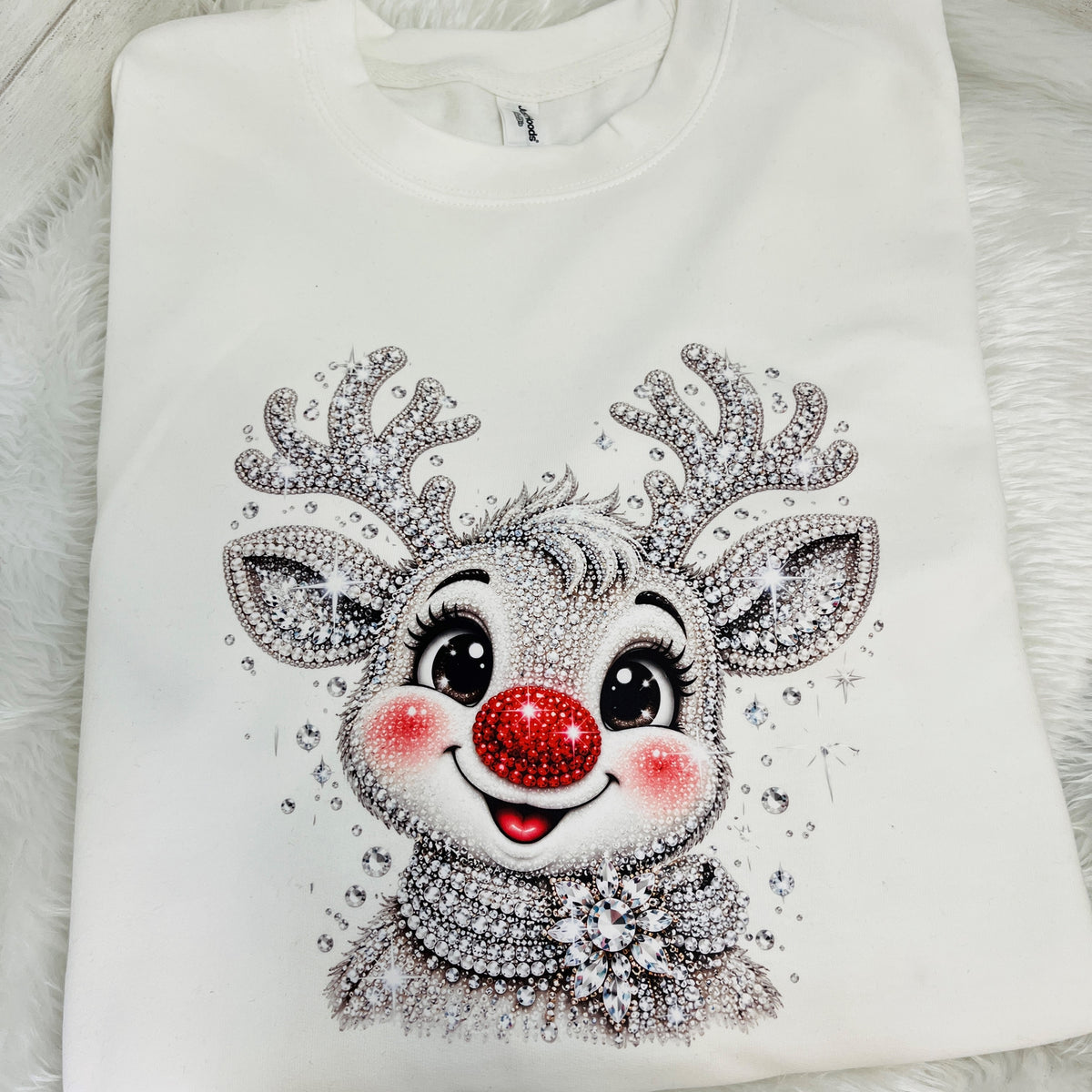 A white long-sleeve sweatshirt featuring a large, detailed graphic of a reindeer face embellished with silver rhinestones, a red rhinestone nose, and pink rhinestone cheeks, laid flat on a white faux fur background.