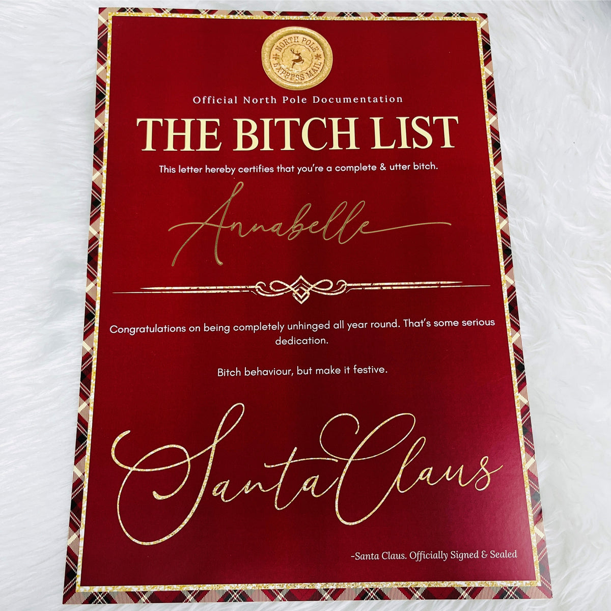 A rectangular red novelty letter or certificate with gold text reading 'THE BITCH LIST' and 'Santa Claus,' plus a gold crest at the top, photographed on a white textured fabric background.