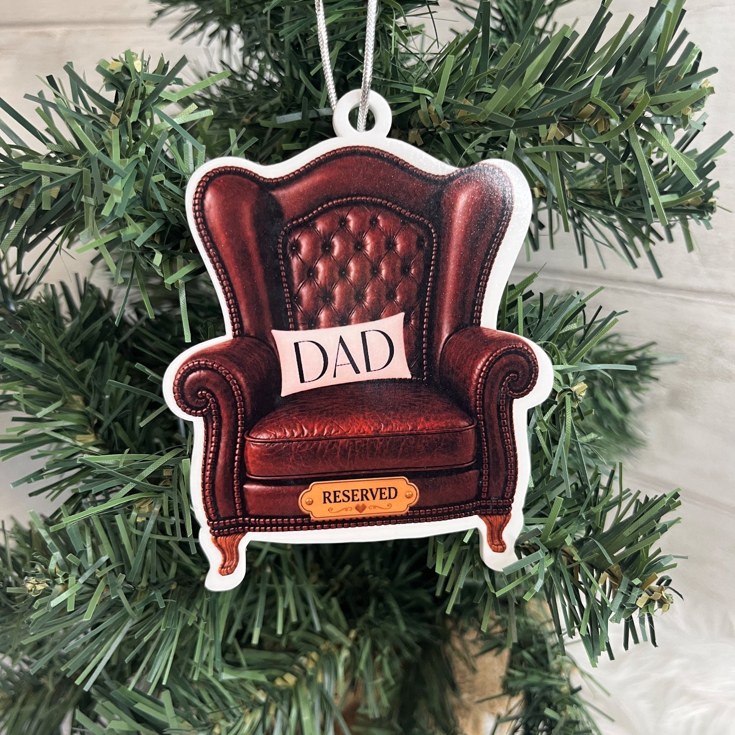 A shaped Christmas ornament depicting a brown leather armchair with a cushion that says 'DAD' and a small sign reading 'RESERVED', hanging on a green Christmas tree.