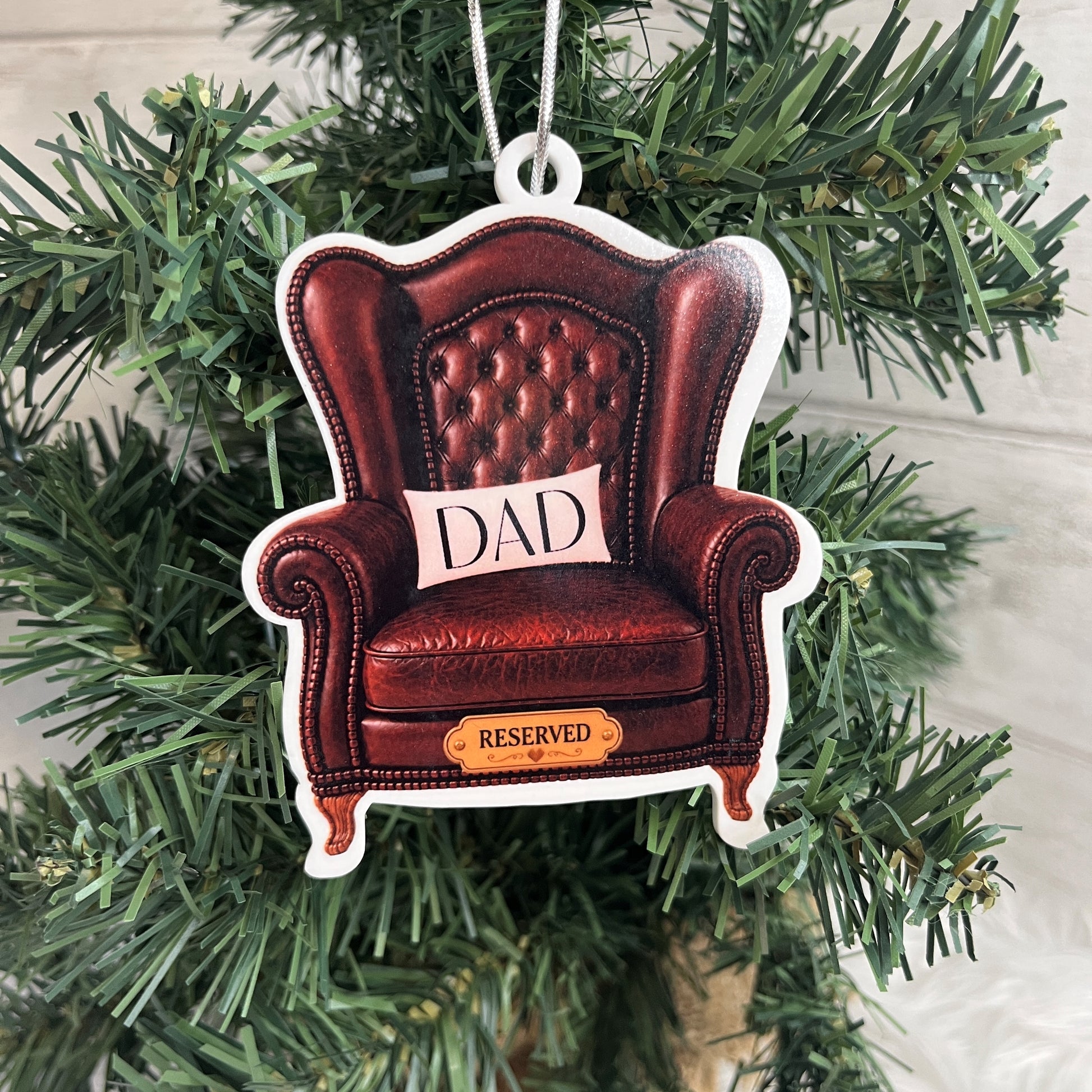 A shaped Christmas ornament depicting a brown leather armchair with a cushion that says 'DAD' and a small sign reading 'RESERVED', hanging on a green Christmas tree.