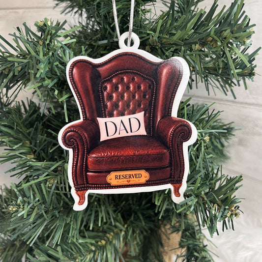 A shaped Christmas ornament depicting a brown leather armchair with a cushion that says 'DAD' and a small sign reading 'RESERVED', hanging on a green Christmas tree.