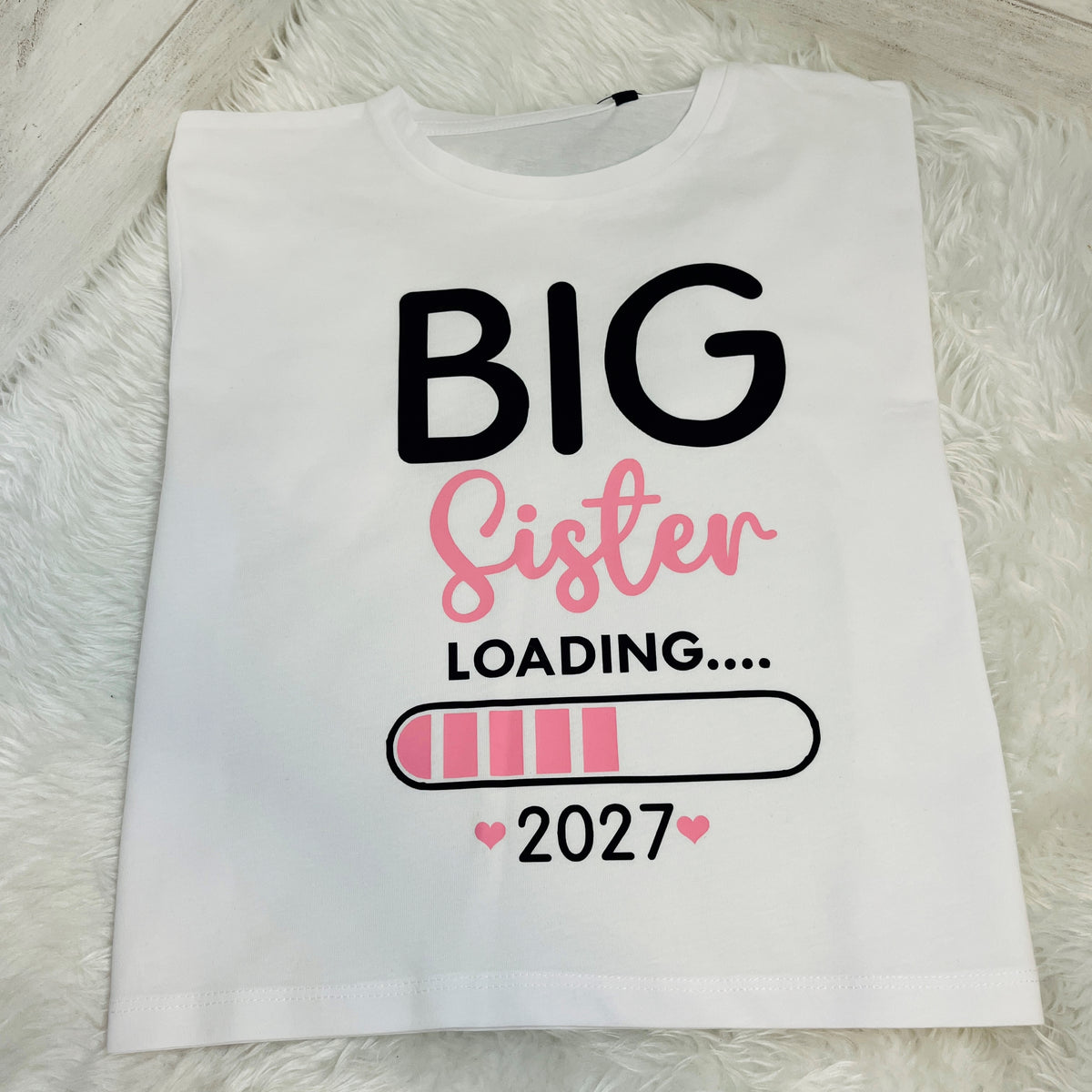 A white short-sleeve T-shirt laid flat on a white faux-fur surface, featuring the text 'BIG Sister LOADING.