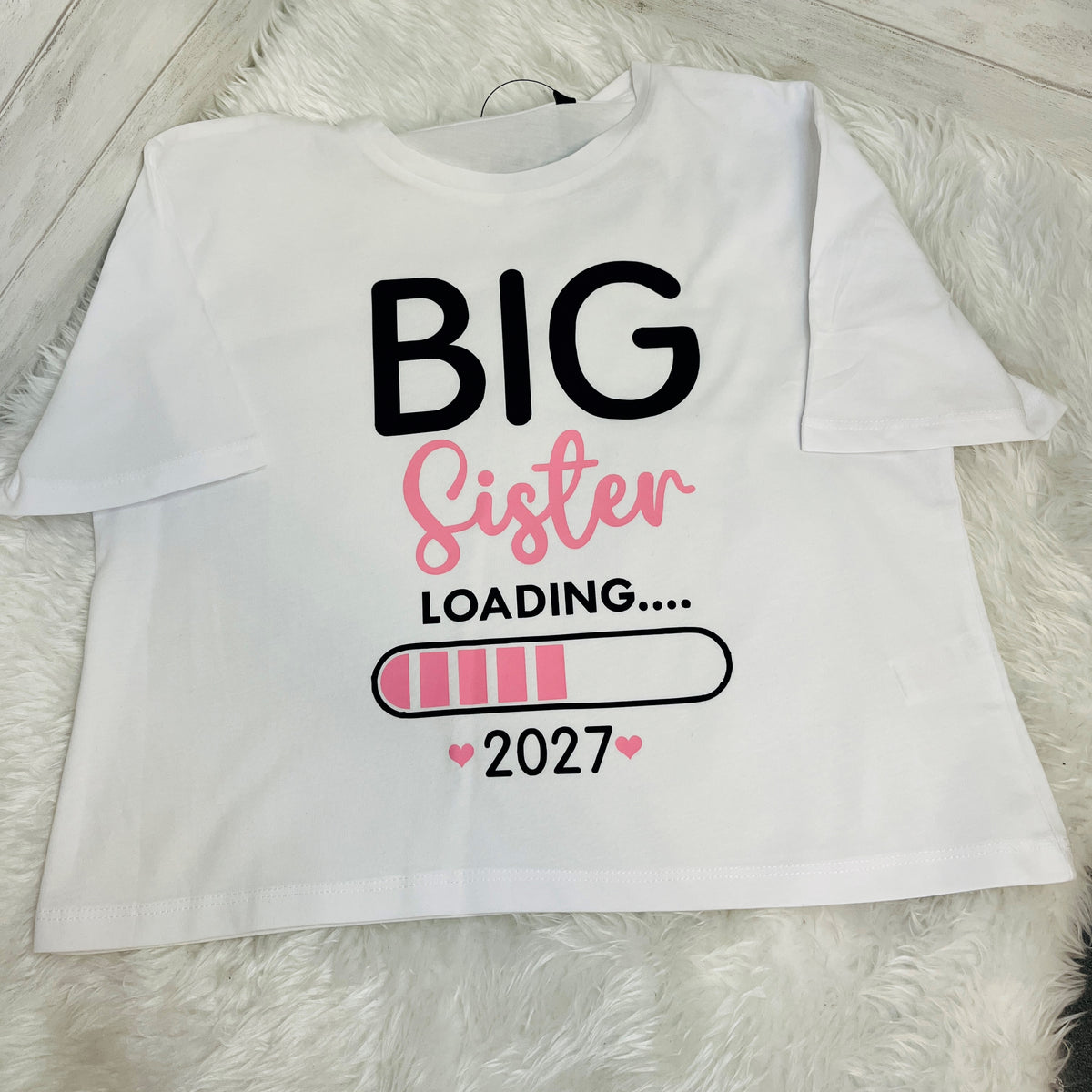 A white short-sleeve t-shirt laid flat on a white faux fur rug, featuring a graphic print that reads 'BIG Sister LOADING.