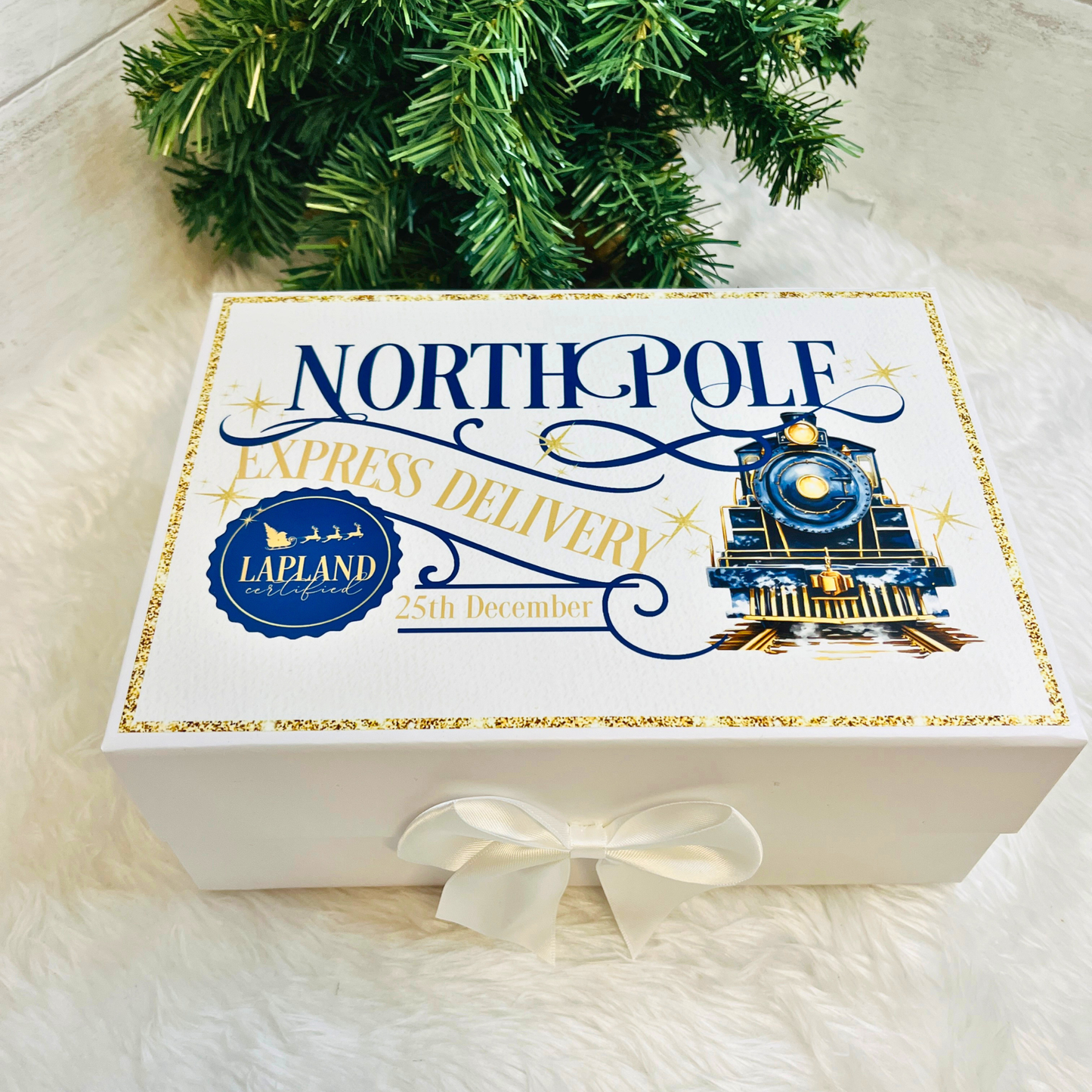 A white rectangular gift box with a bow on the front, printed with 'NORTH POLE EXPRESS DELIVERY 25th December' and a blue train illustration, styled on a white furry surface with green pine branches behind it.