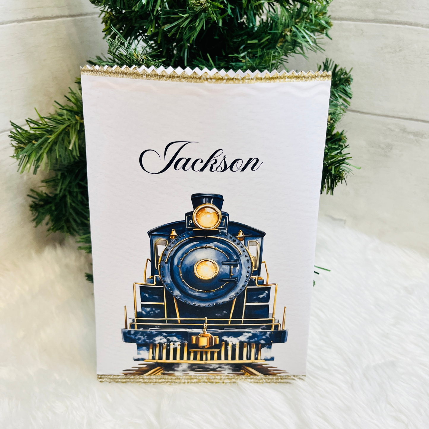 A personalized white candy bag featuring a navy blue and gold steam locomotive illustration and the name 'Jackson' in navy script, styled with green pine branches on a white faux fur surface.