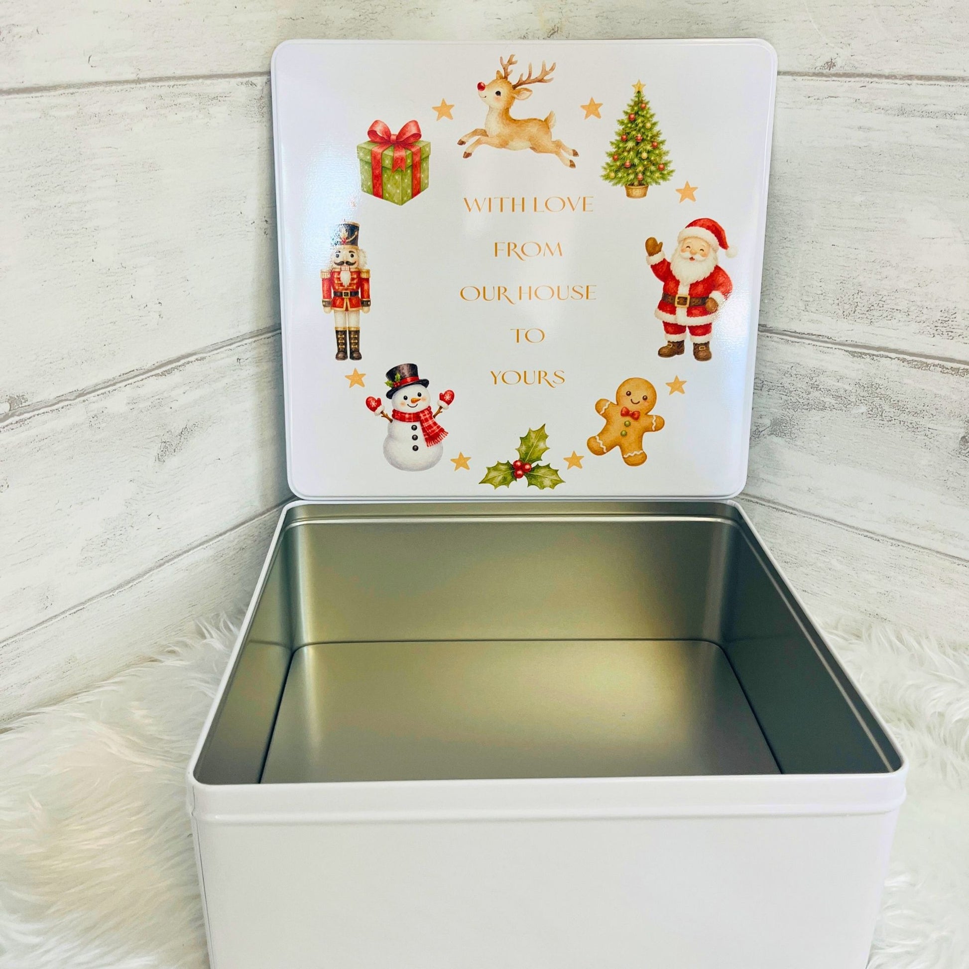 A square white metal gift tin with its lid open, showing a festive Christmas-themed printed design on the inside lid with illustrations of Santa Claus, a snowman, a nutcracker, a reindeer, a gingerbread man, and a gift box, plus the text 'WITH LOVE FROM OUR HOUSE TO YOURS'. The tin is photographed on a white faux-fur surface against a light wood plank background.