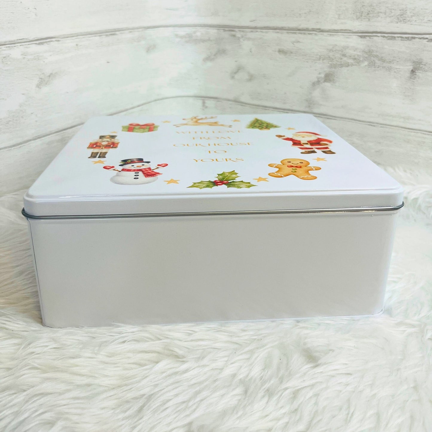 A square white metal gift tin with a Christmas-themed lid illustration featuring a snowman, Santa, a gingerbread man, holly, placed on a white faux fur surface against a white wood-paneled background.