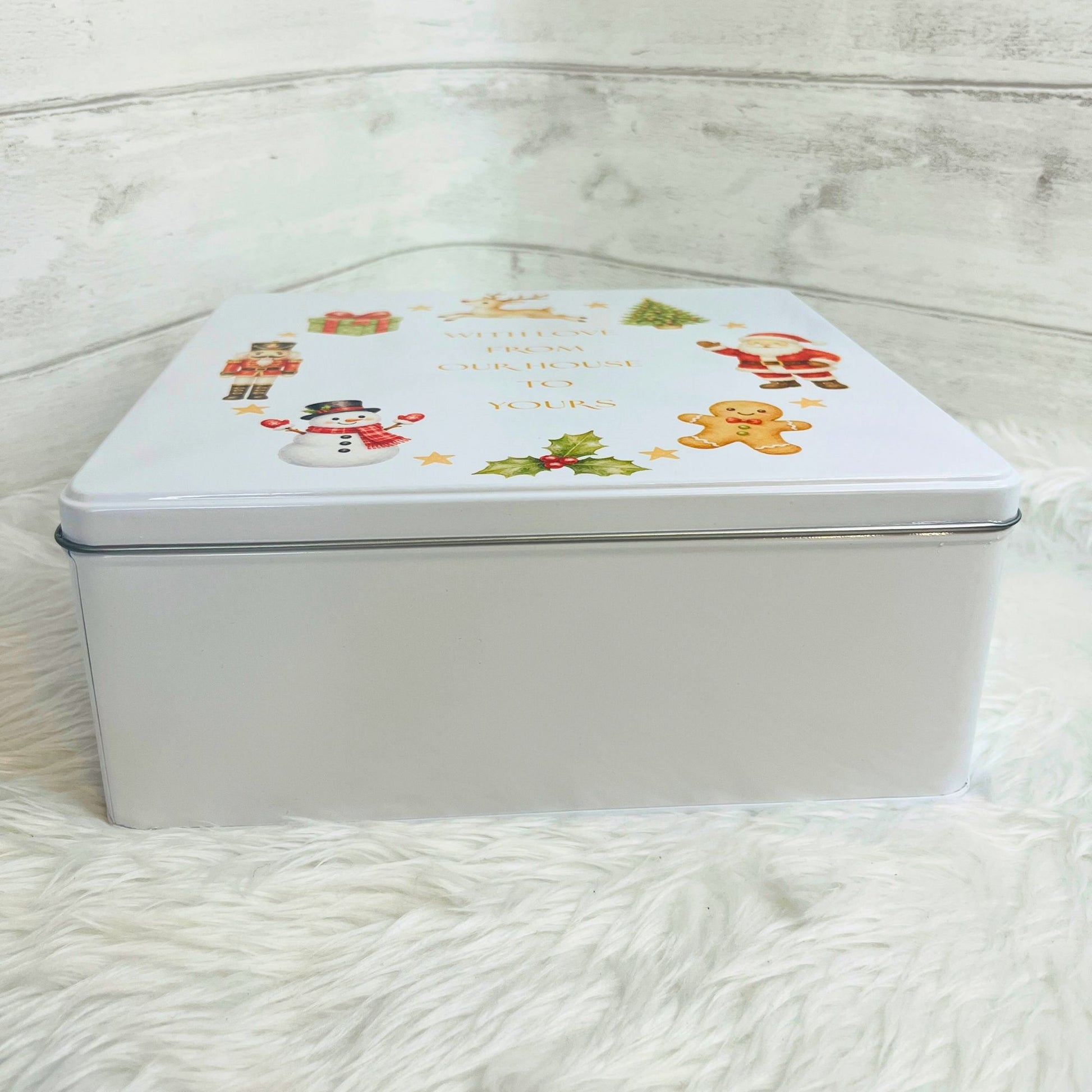 A square white metal gift tin with a Christmas-themed lid illustration featuring a snowman, Santa, a gingerbread man, holly, placed on a white faux fur surface against a white wood-paneled background.