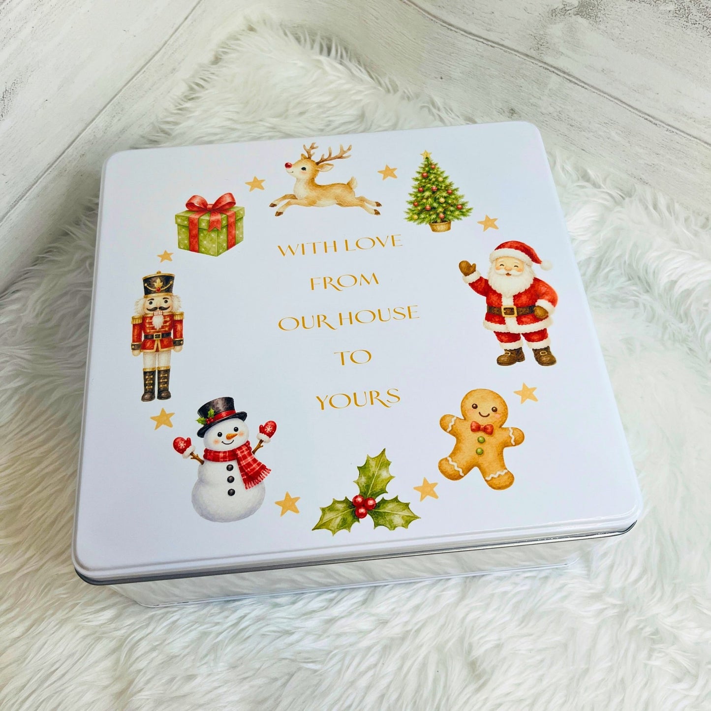 A square white Christmas-themed metal tin with a printed holiday design, placed on a white faux-fur surface. The tin features illustrations of Santa Claus, a snowman, a nutcracker, a gingerbread man, a Christmas tree, a gift box, a reindeer, holly, and gold text reading 'WITH LOVE FROM OUR HOUSE TO YOURS'.
