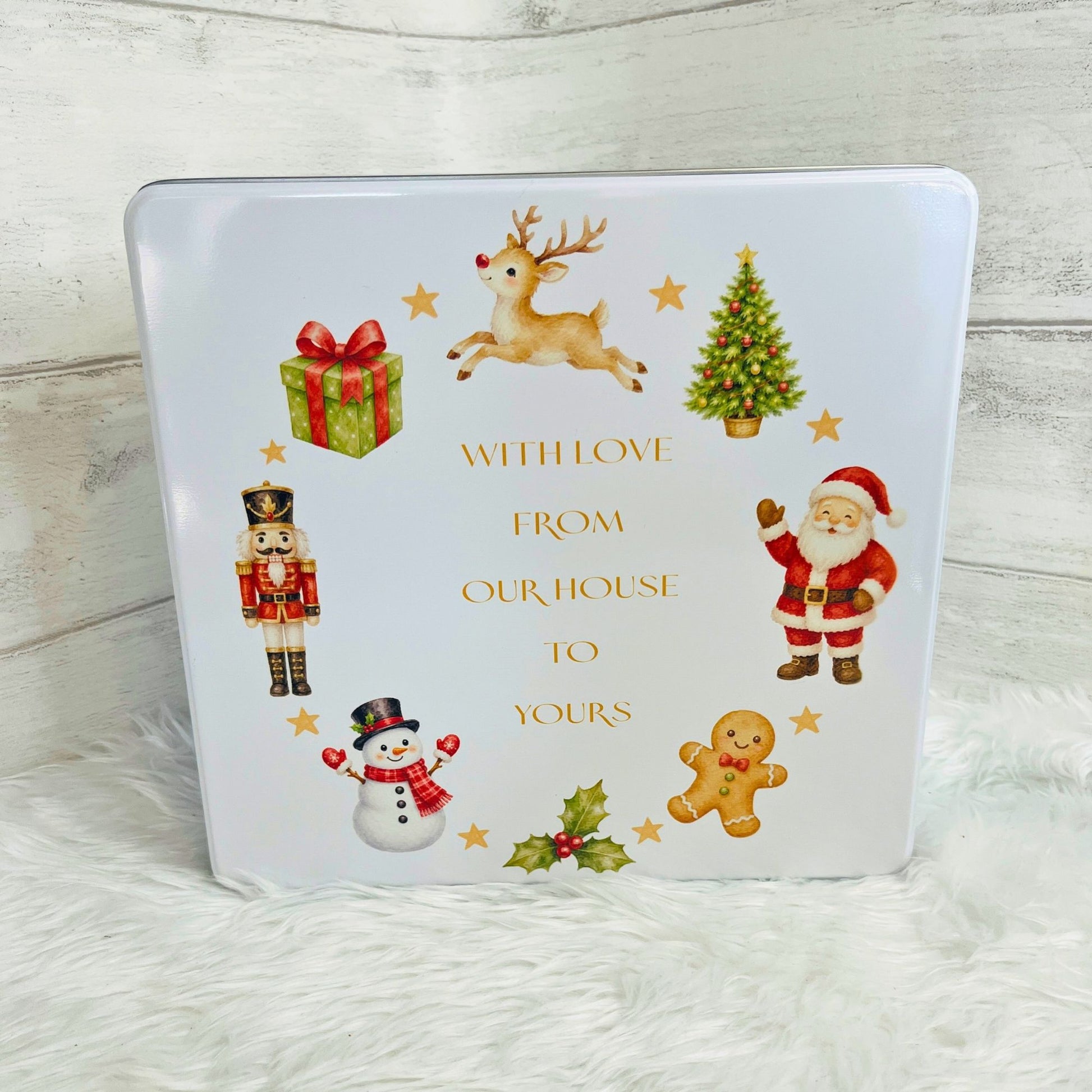A square white Christmas-themed keepsake box or tin with festive illustrations including Santa Claus, a reindeer, a nutcracker, a snowman, a gingerbread man, a gift box, a Christmas tree, holly, and gold text reading 'WITH LOVE FROM OUR HOUSE TO YOURS', shown on a white faux-fur surface against a light wood background.