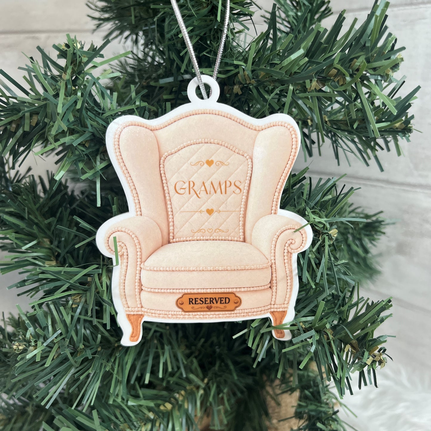 A decorative ornament shaped like a pale cream wingback armchair with the word “GRAMPS” stitched on the backrest and a small “RESERVED” plaque on the front, hanging from a silver string among green Christmas tree branches.