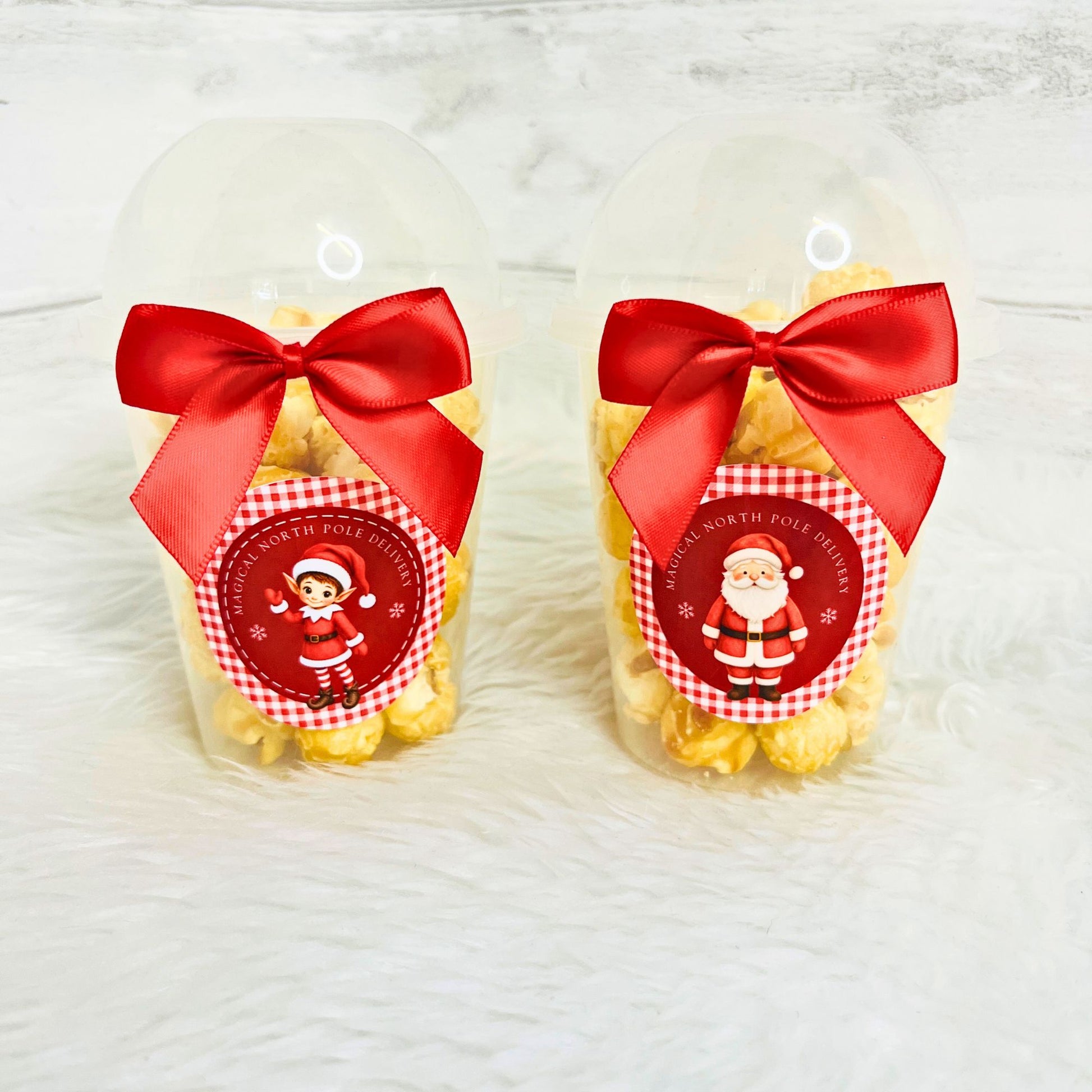 Two clear plastic dome cups filled with yellow popcorn, tied with large red satin bows and decorated with round Santa-themed labels, set on a white faux-fur surface.