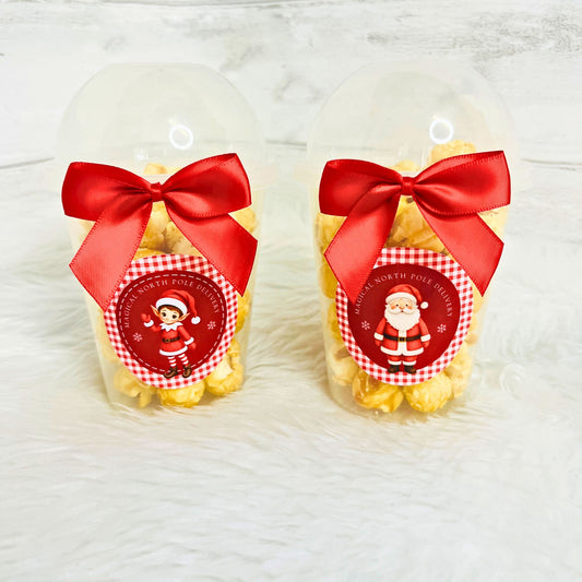 Two clear plastic dome cups filled with yellow popcorn, tied with large red satin bows and decorated with round Santa-themed labels, set on a white faux-fur surface.