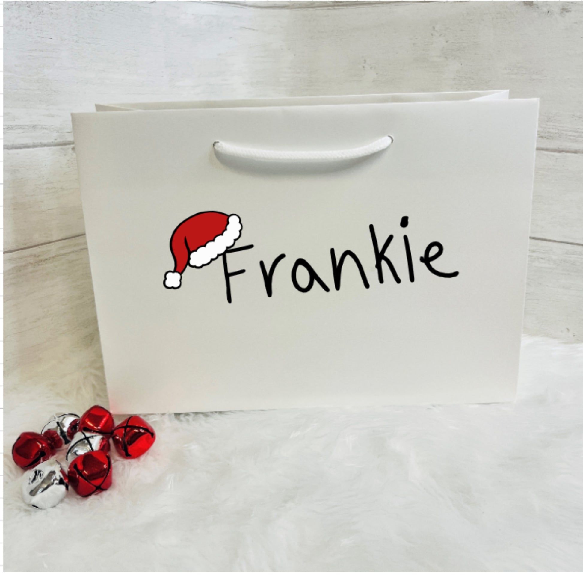 White gift bag with 'Frankie' and a Santa hat design, on a light wooden surface.