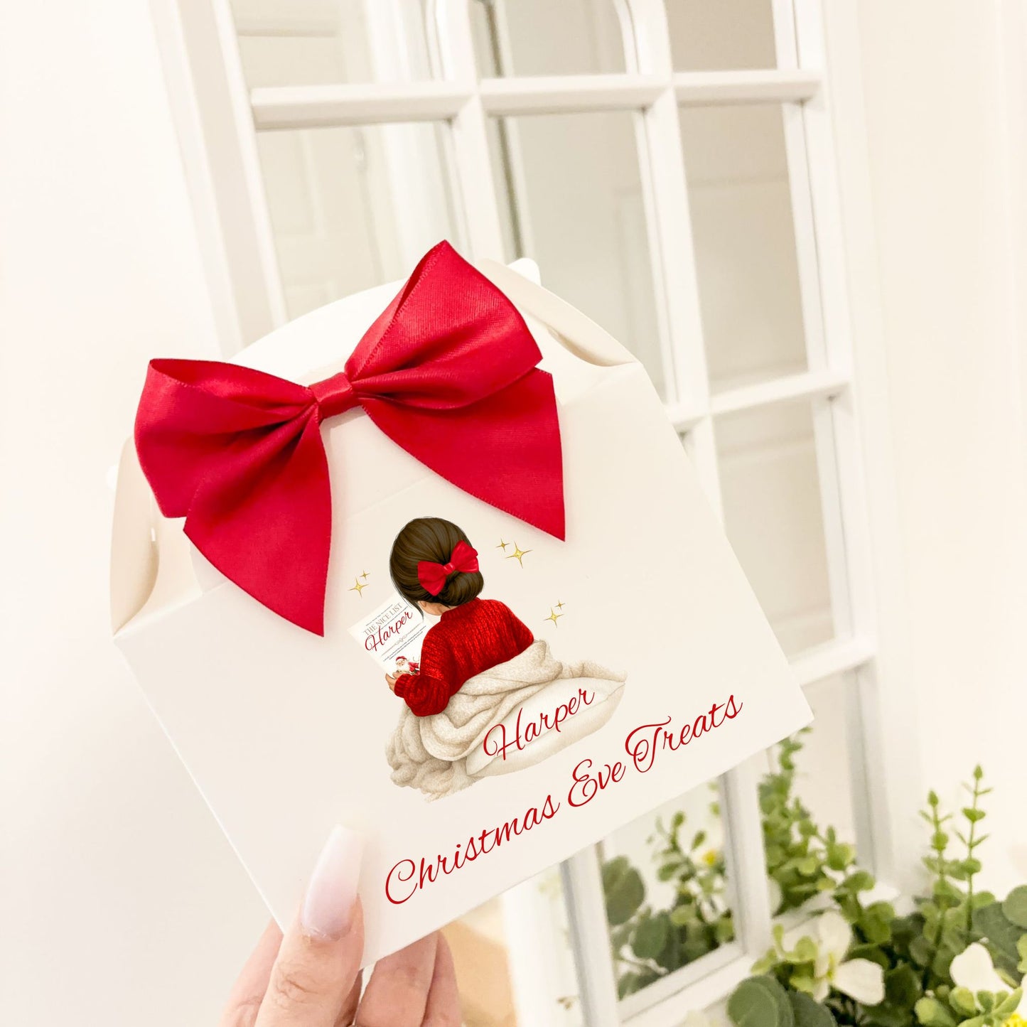 A hand holding a white Christmas Eve treats gift box decorated with a large red satin bow, featuring an illustration of a girl in a red sweater and the text 'Harper's Christmas Eve Treats'.