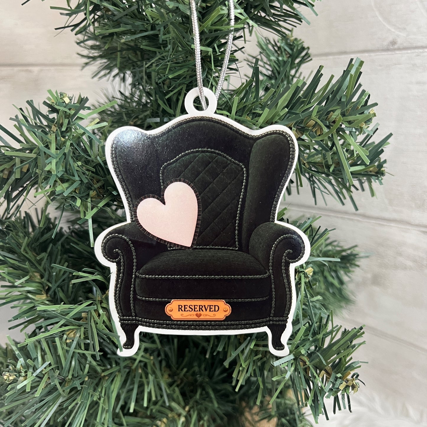 A dark green wingback armchair Christmas ornament with a quilted pattern, a light pink heart on the back, and a small gold 'RESERVED' plaque at the bottom, hanging on a green Christmas tree.