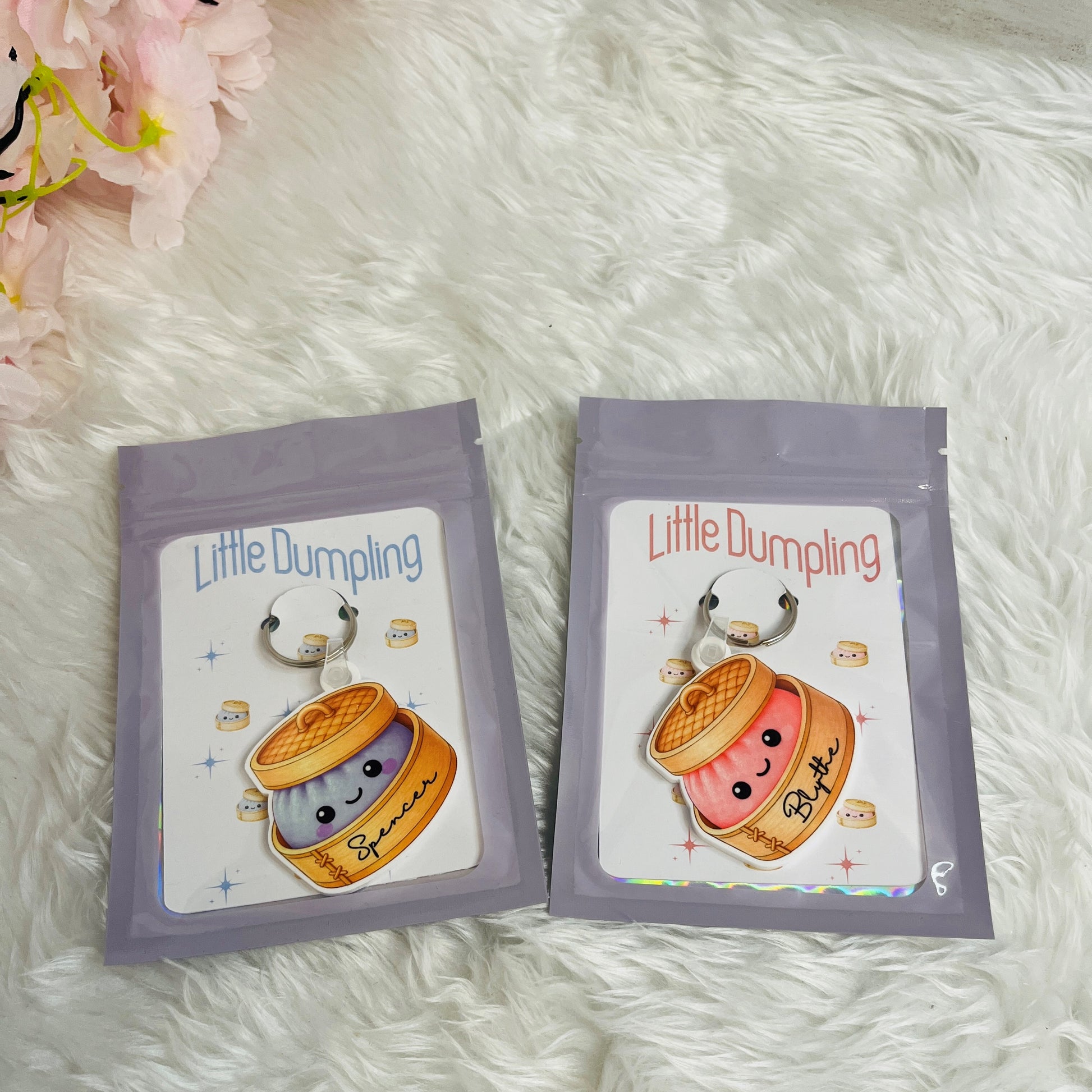 Two packages of 'Little Dumpling' keyrings on a backing card in a plastic wallet on a fluffy white surface with flowers in the corner.