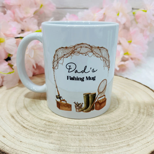 White mug with 'Dad's Fishing Mug' text and fishing-themed illustrations on a wooden surface 
