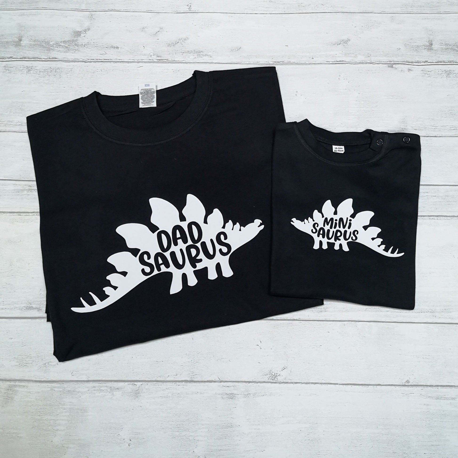 Two black t-shirts with dinosaur-themed text on a light wooden surface