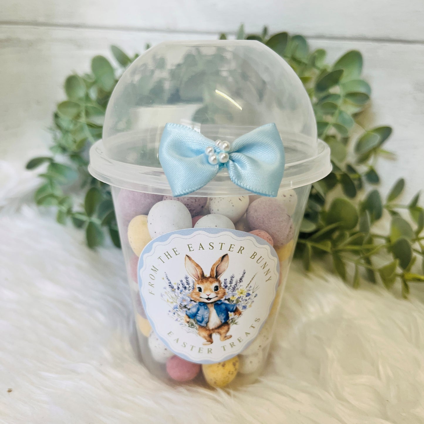 An Easter Dome Snack Pot featuring a plastic cup with a blue bunny wreath design and a blue bow on the lid, filled with various small snacks.