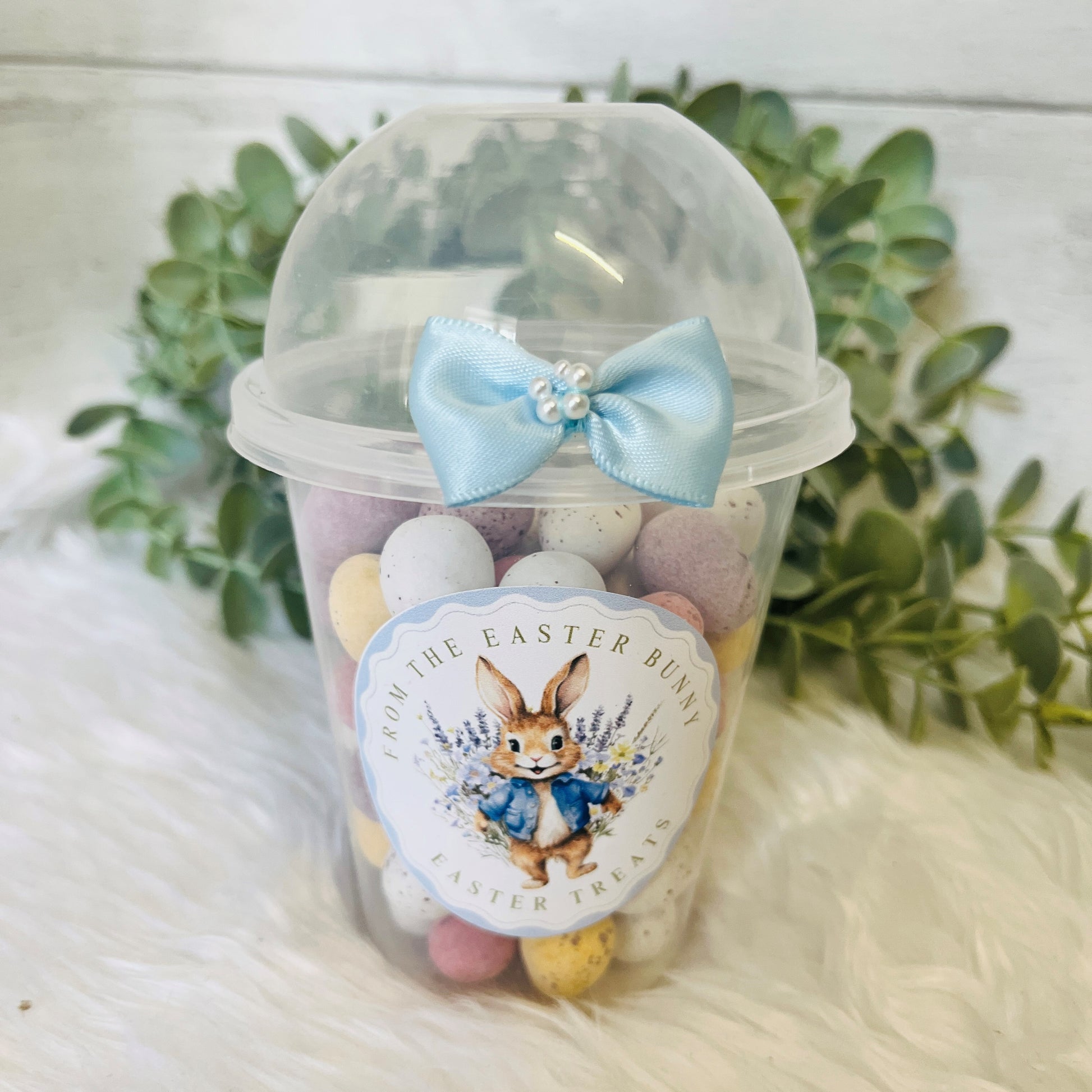 An Easter Dome Snack Pot featuring a plastic cup with a blue bunny wreath design and a blue bow on the lid, filled with various small snacks.