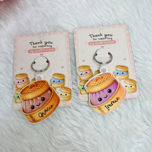 Two cute cartoon keychains shaped like smiling dumplings on pink backing cards, placed on a white faux-fur surface. 