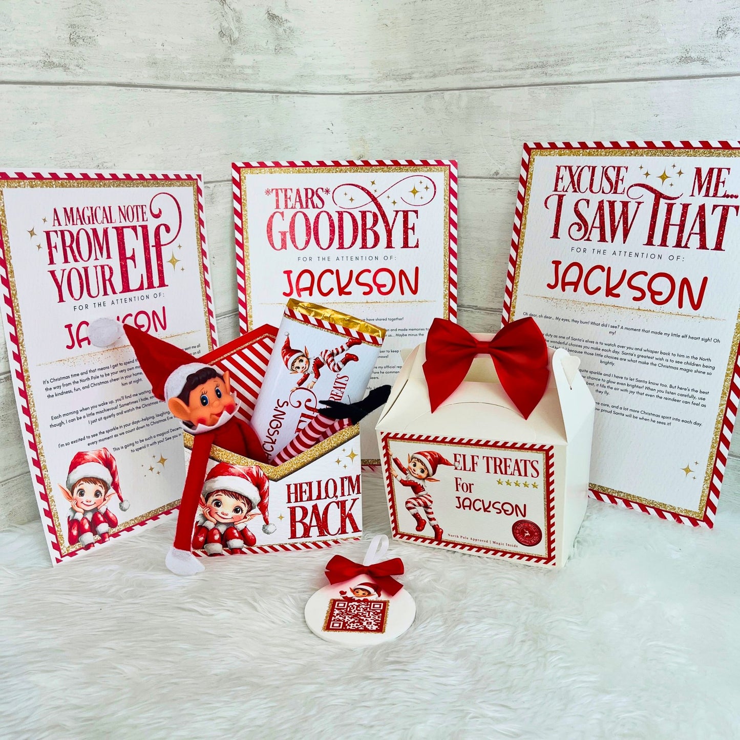 A Christmas-themed elf gift set displayed on a white faux fur surface against a light wood plank background. The set includes three printed holiday message cards addressed to Jackson, a small white gift box with a red bow, an elf treat bag, an 8cm acrylic santa tracker, and a small elf figure