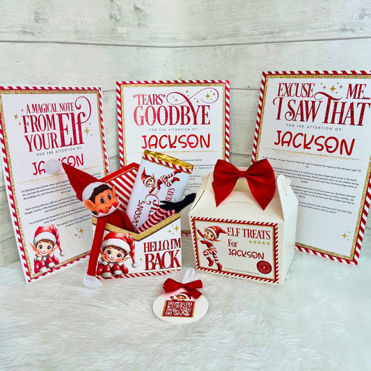 A Christmas-themed elf gift set displayed on a white faux fur surface against a light wood plank background. The set includes three printed holiday message cards addressed to Jackson, a small white gift box with a red bow, an elf treat bag, an 8cm acrylic santa tracker, and a small elf figure