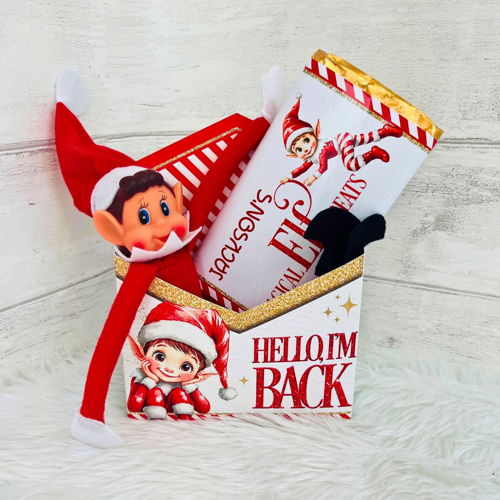 A personalized Elf on the Shelf gift set featuring a plush Elf on the Shelf toy, a red Santa hat, and a chocolate bar wrapped in a white paper sleeve printed with 'Jackson's Magical Elf Treats and 'HELLO I'M BACK' with an elf illustration, arranged in a box on a white faux fur surface against a light wood background.