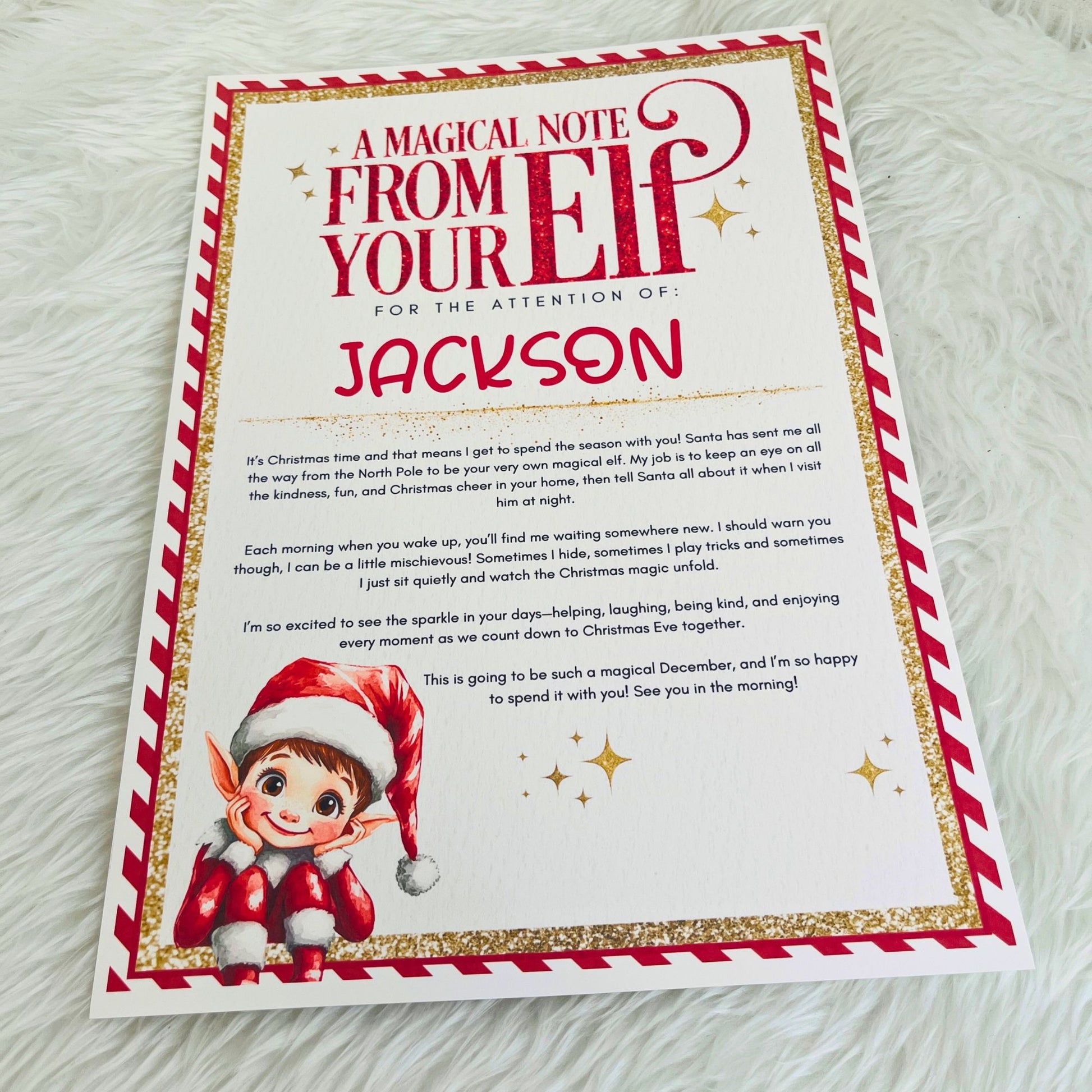 A personalized Christmas letter featuring a festive red and white border, gold glitter accents, and a cute elf illustration at the bottom. The card is printed with a magical note to 'Jackson' and is photographed flat on a soft white faux fur background.