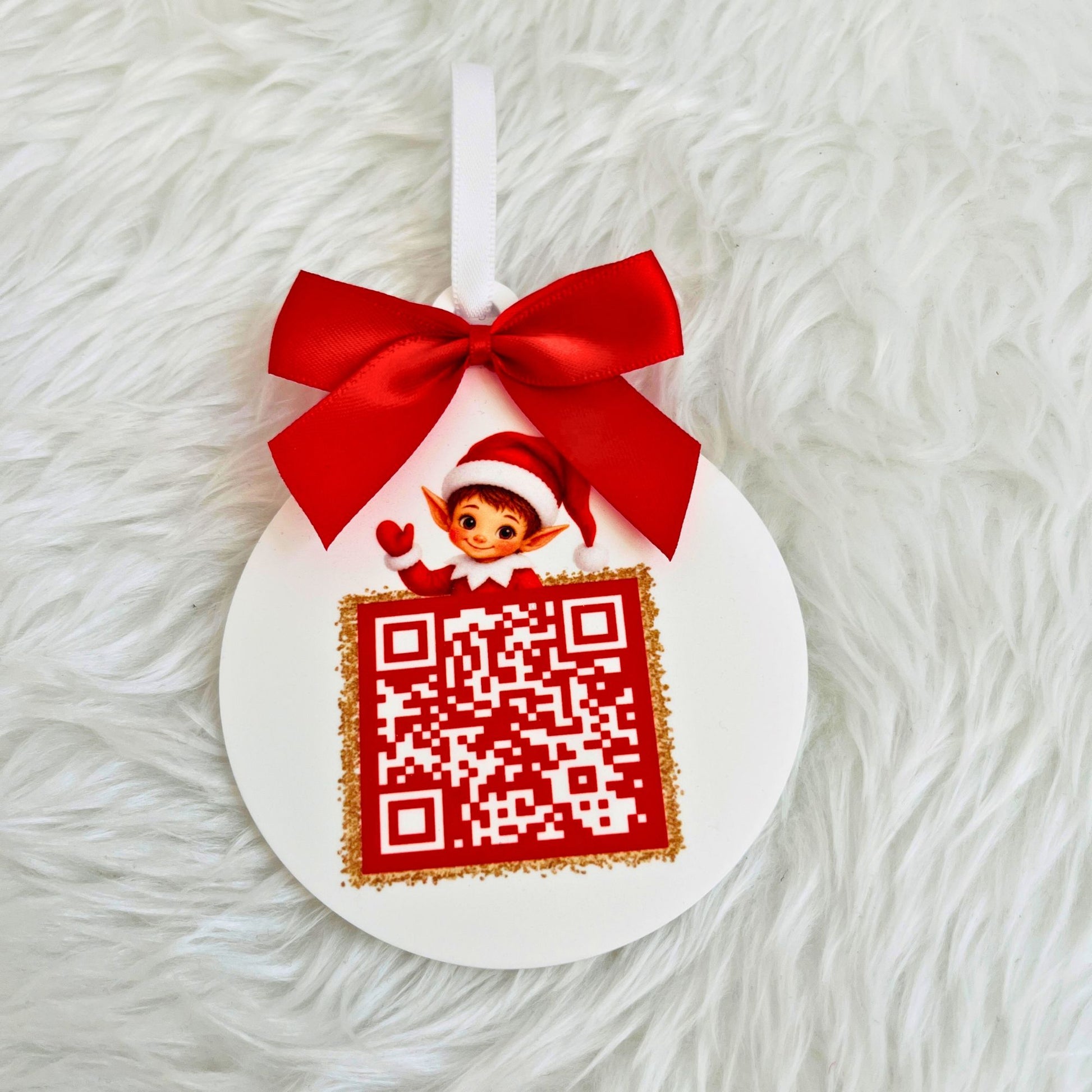 A round 8cm white acrylic Santa Tracker ornament with a red ribbon bow at the top, featuring a small elf illustration above a red QR code with gold glitter trim, photographed on a white faux-fur background.