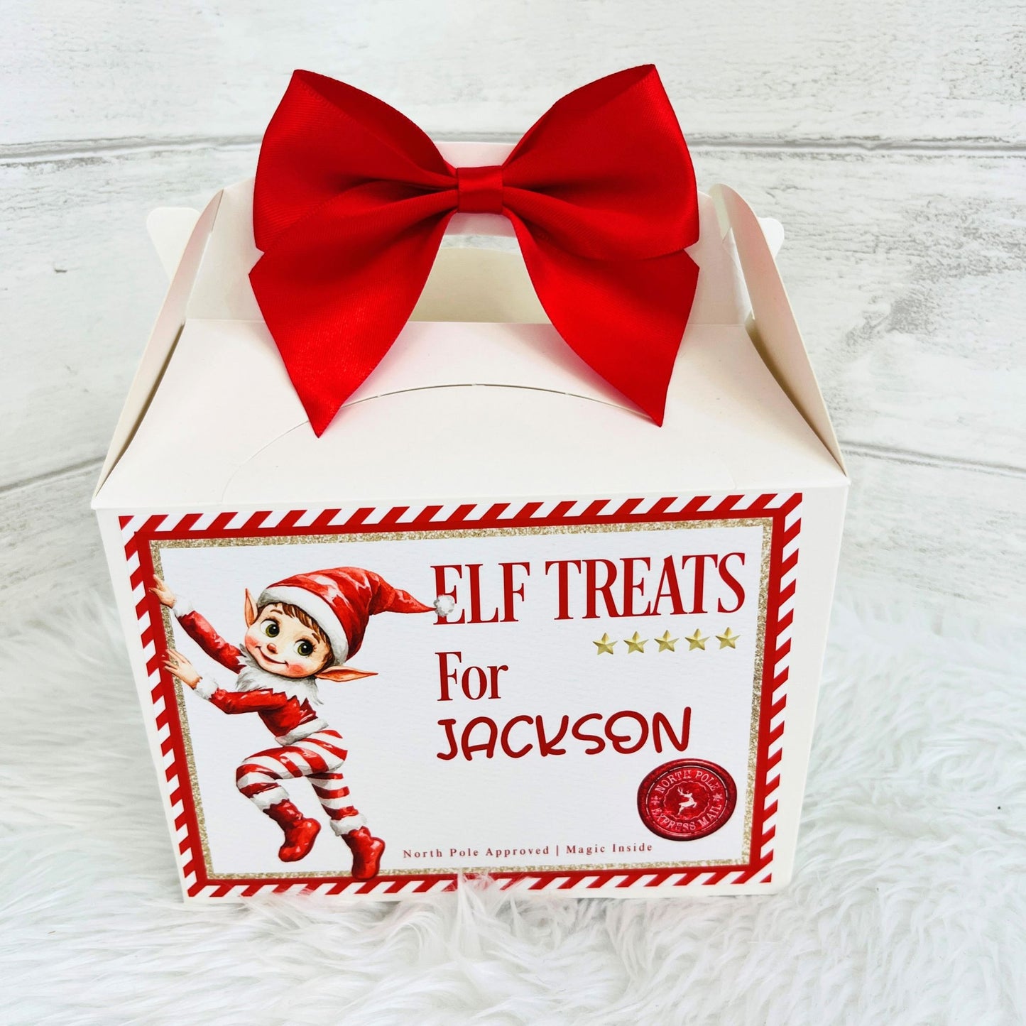 A white gift box with a large red bow on top, featuring a front label that reads 'ELF TREATS For Jackson' with an elf illustration and a red circular seal. The box is photographed on a white faux-fur surface against a light wood background.
